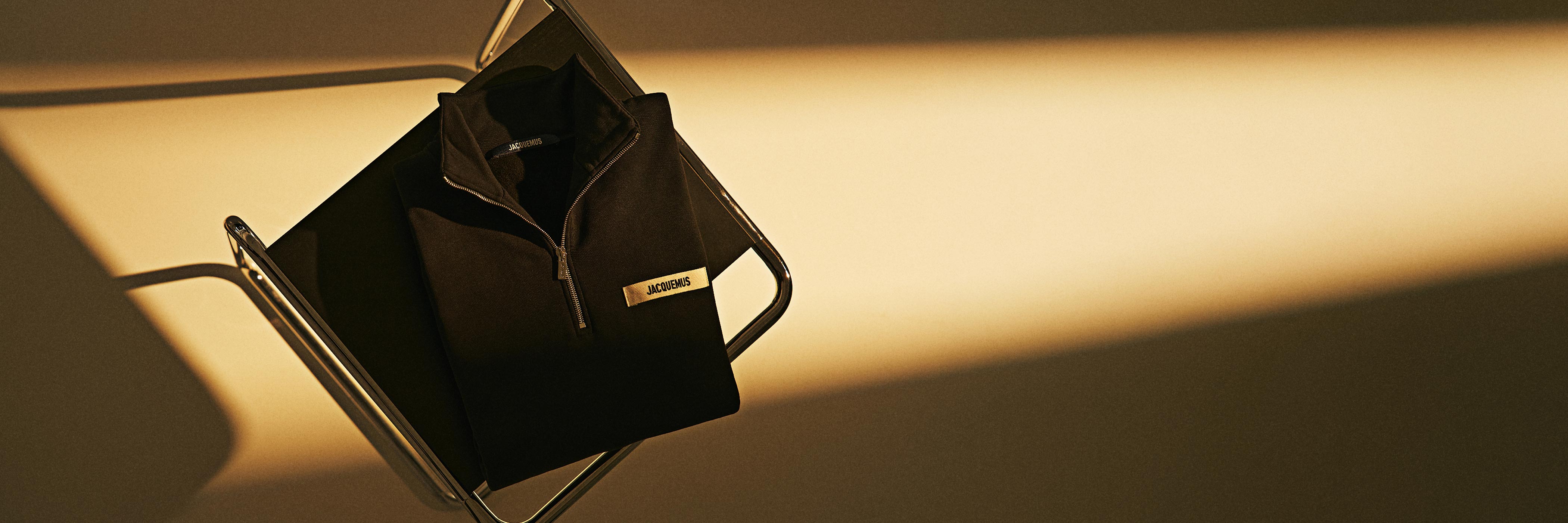 Schwarzer Jacquemus Pullover mit Reißverschluss liegt gefaltet auf modernem schwarzem Stuhl im warmen Licht mit Schatten.
