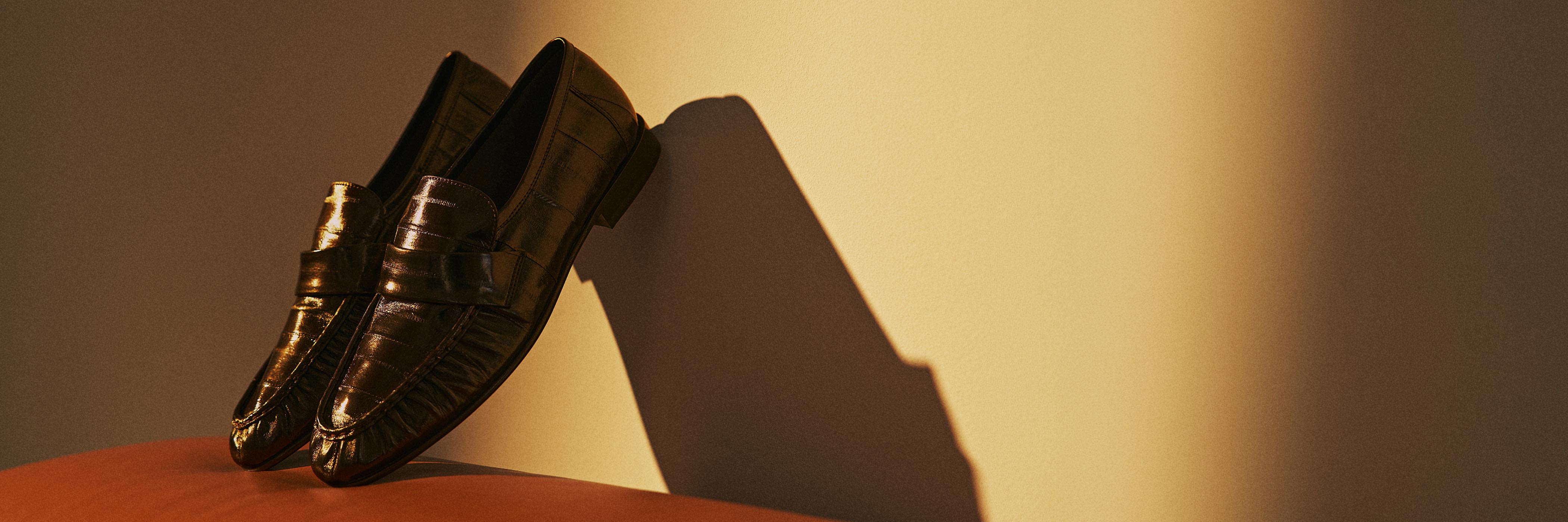 Elegante braune Lederschuhe lehnen an einer Wand mit warmem Licht und werfen einen langen Schatten auf die beige Wand.