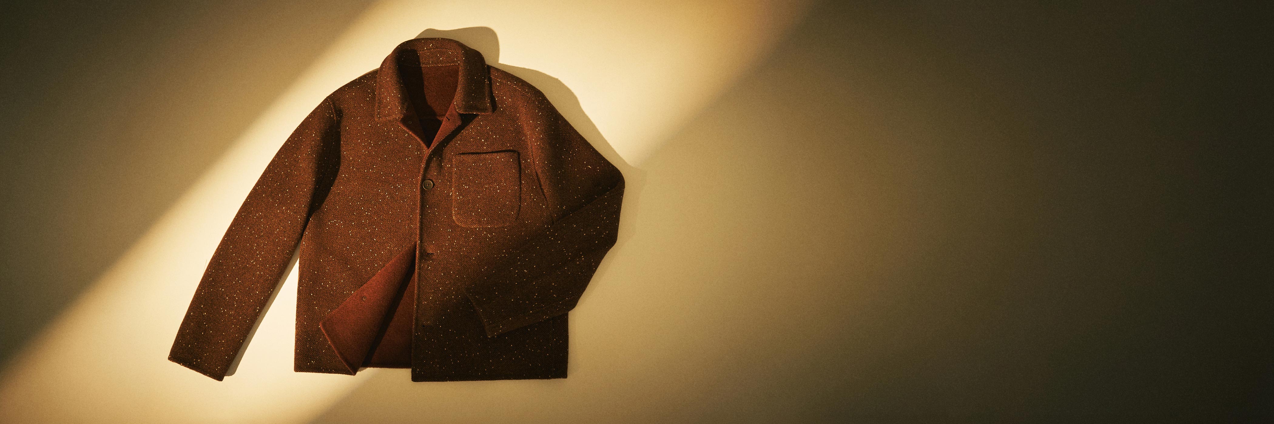 Braune, melierte Jacke mit Kragen und Brusttasche, liegend im warmen Licht auf hellem Hintergrund.