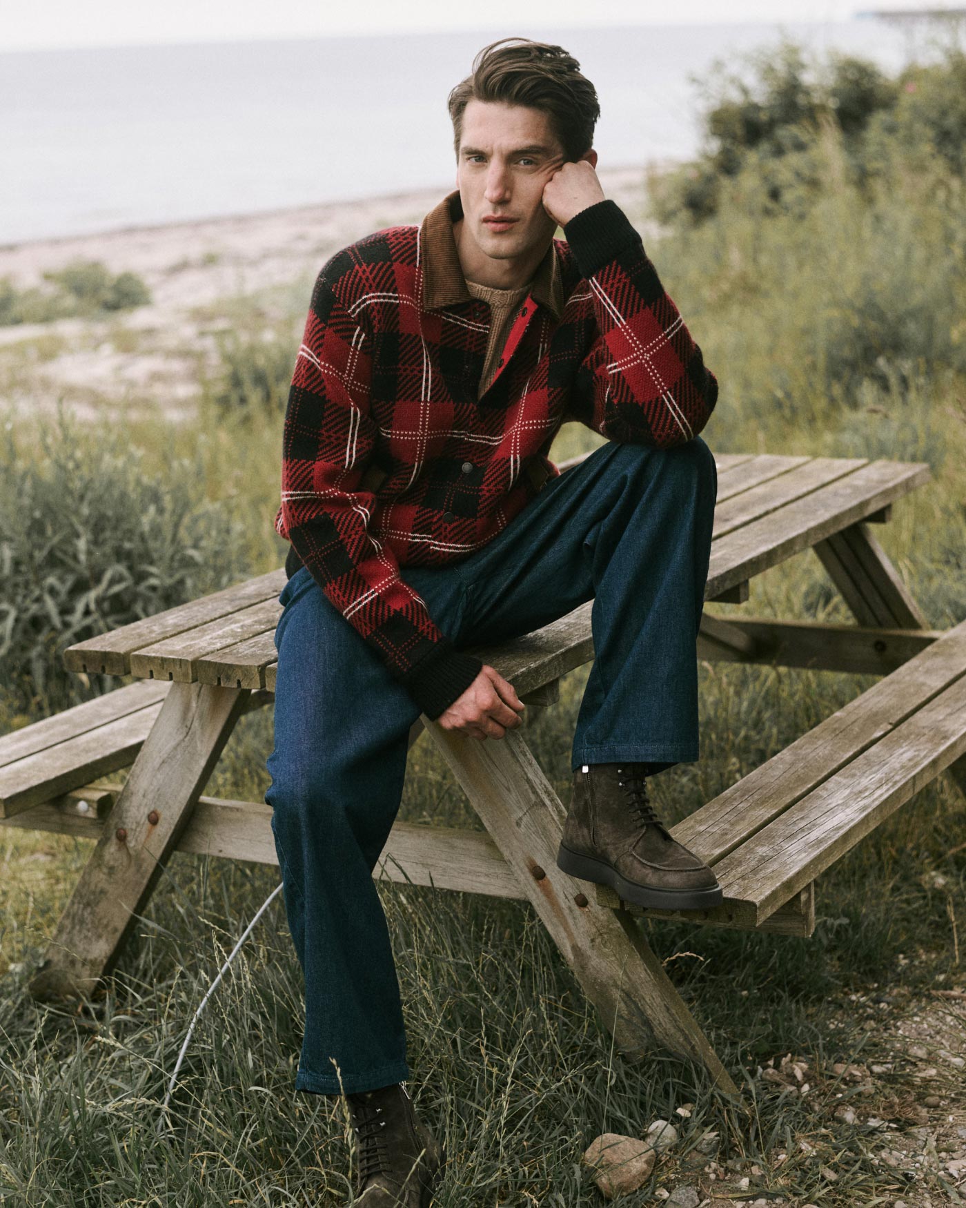 Young man wearing a red and black plaid jacket, blue jeans, and brown boots sitting on a wooden picnic table outdoors.