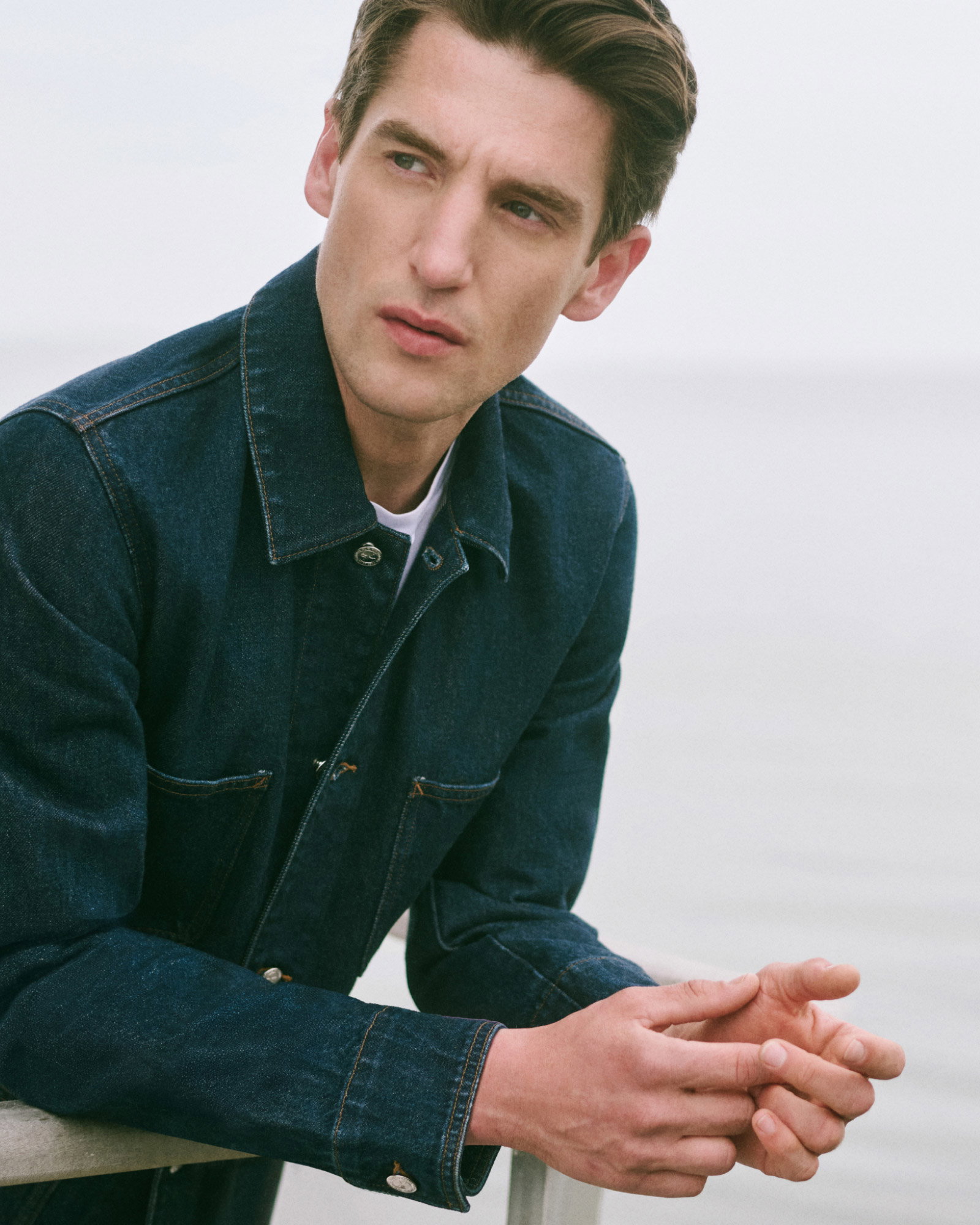 Close-up of a serious young man wearing a dark denim jacket, leaning on a railing with a blurred light background.