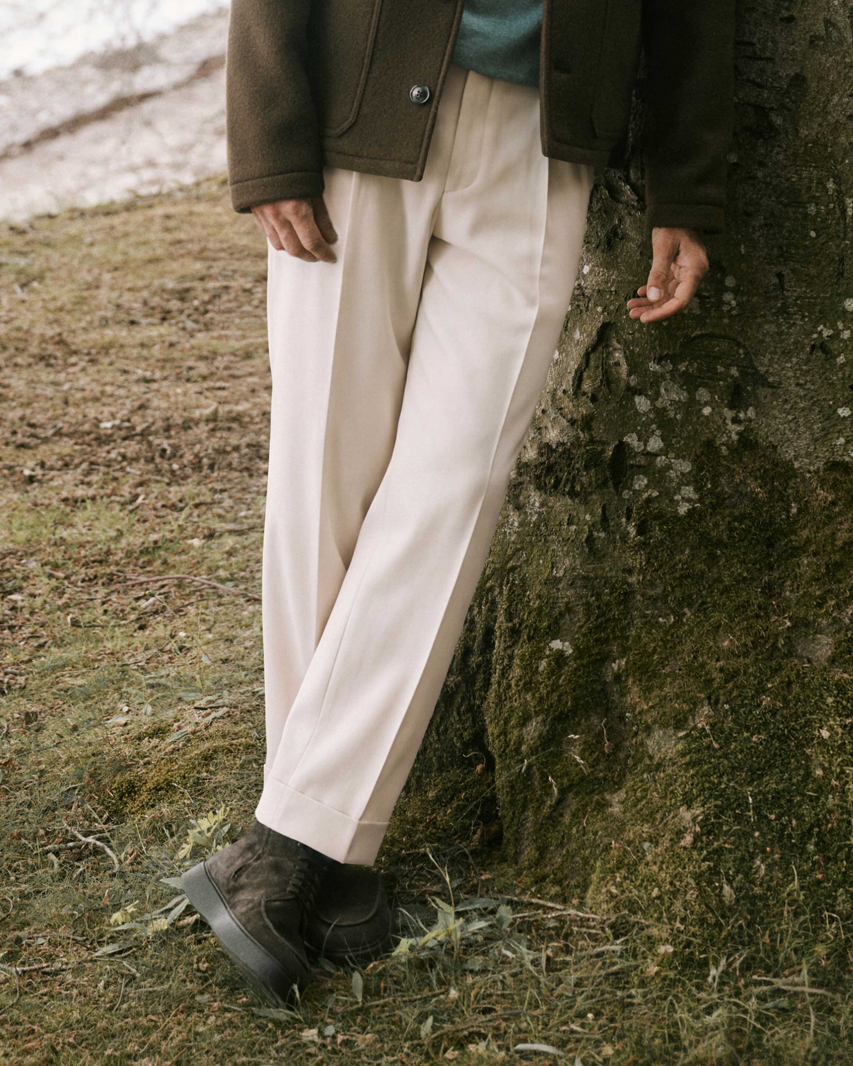 Mann lehnt an Baum, trägt beige weite Hose, dunkelgrünen Wollmantel, hellgrünen Pullover und schwarze Wildlederstiefel.