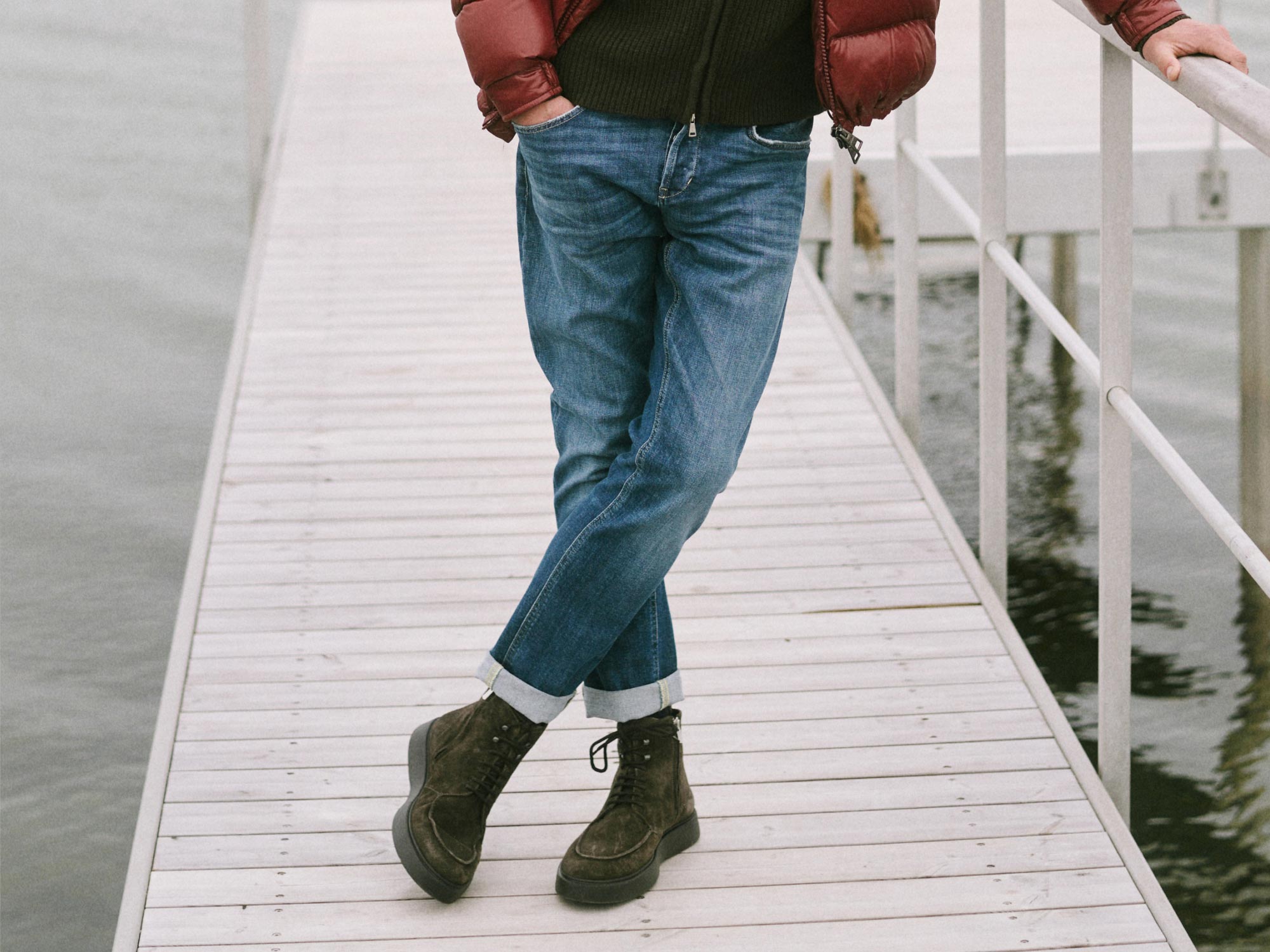Man wearing blue jeans, brown boots, black sweater, and red puffer jacket leaning on a white railing over water on a wooden dock.