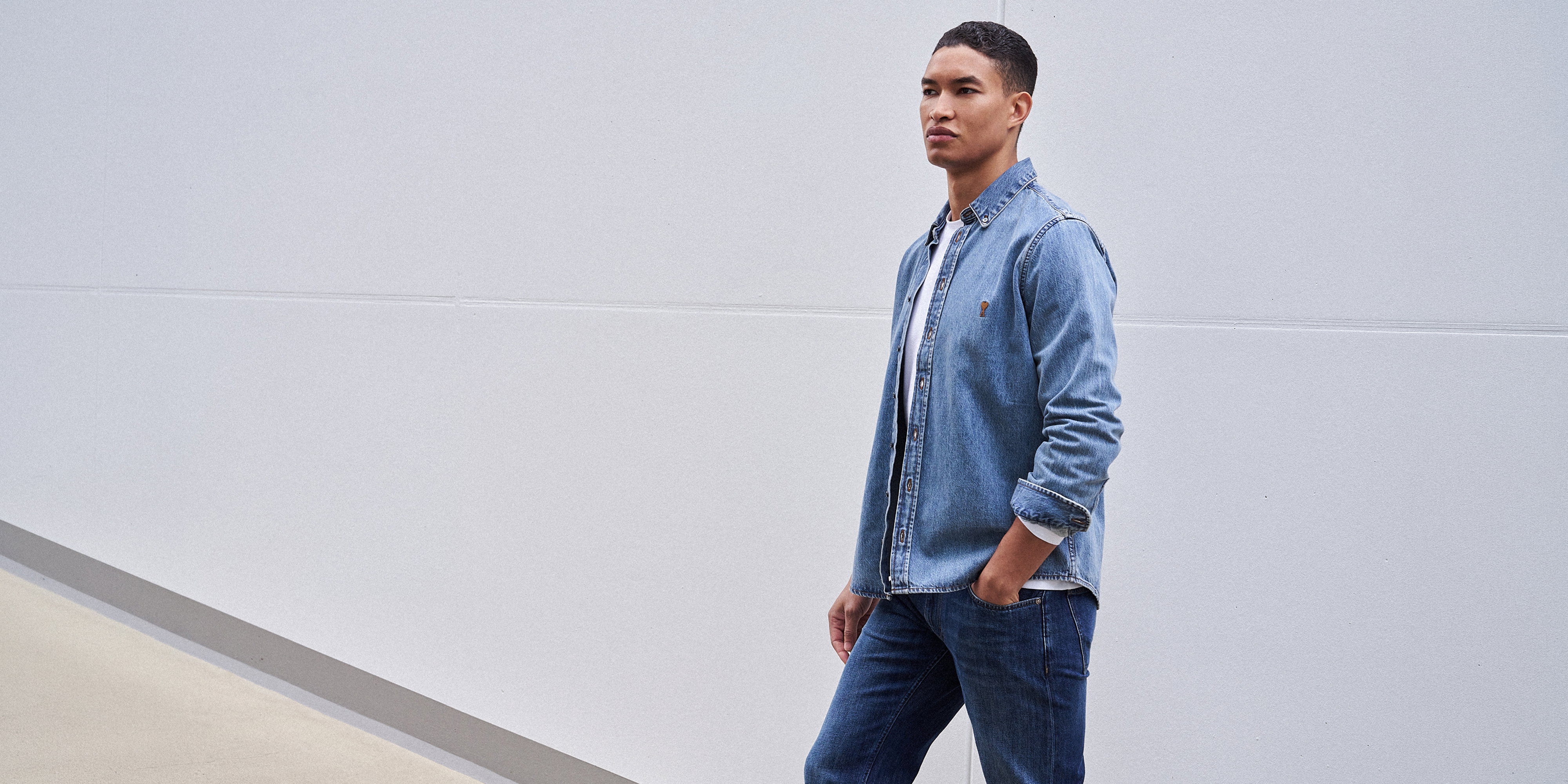 Junger Mann mit dunklen Haaren trägt hellblaue Jeansjacke, weiße T-Shirt, blaue Jeans, steht vor heller Wand, Hand in Tasche.