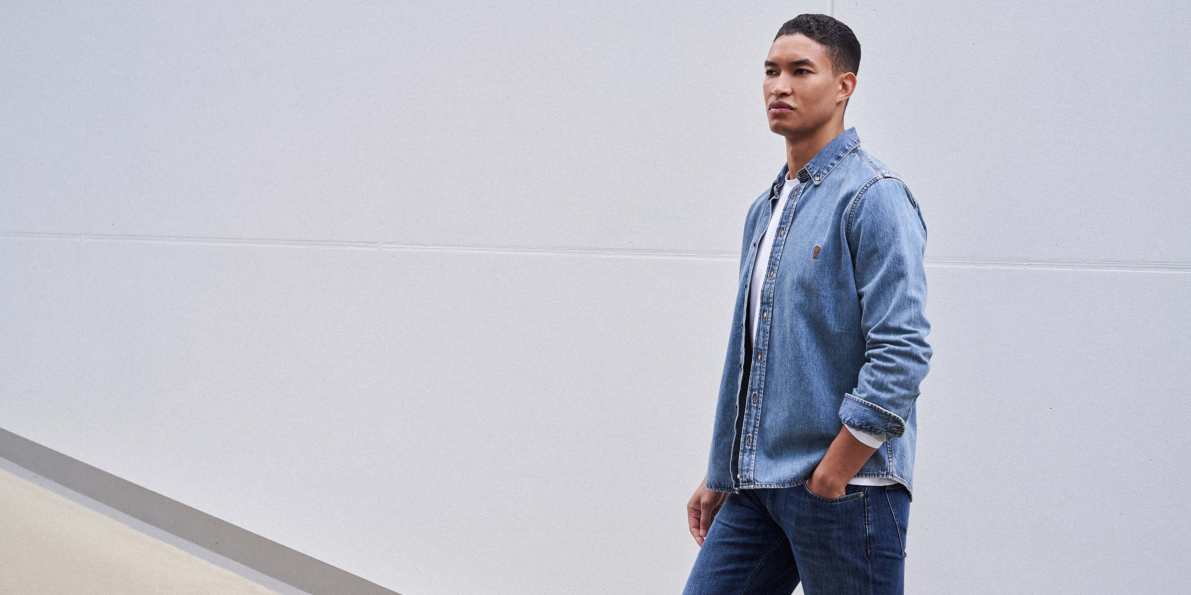 Junger Mann trägt hellblaue Jeansjacke über weißem Shirt und blaue Jeans, steht vor hellgrauer Wand, Hände in Taschen.