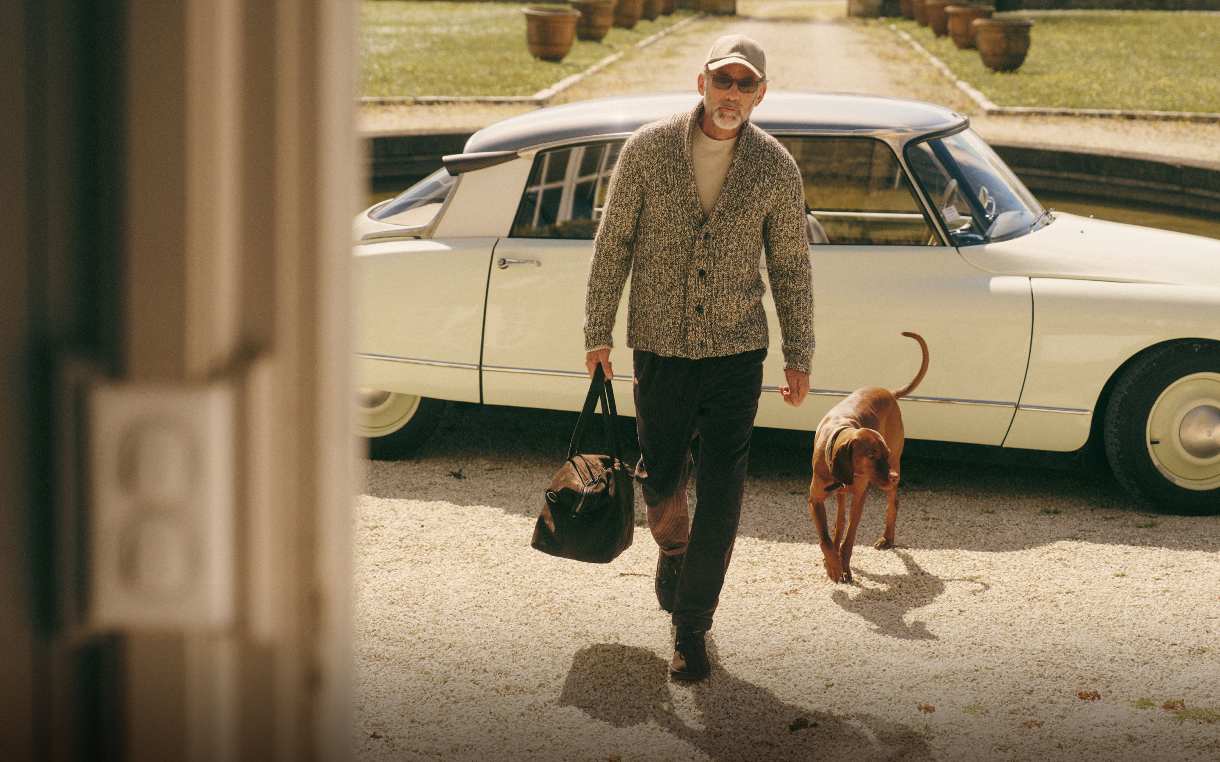 Mann mit grauem Pullover, Cap und Tasche läuft mit braunem Hund vor weißem Oldtimer und Gartenweg entlang.