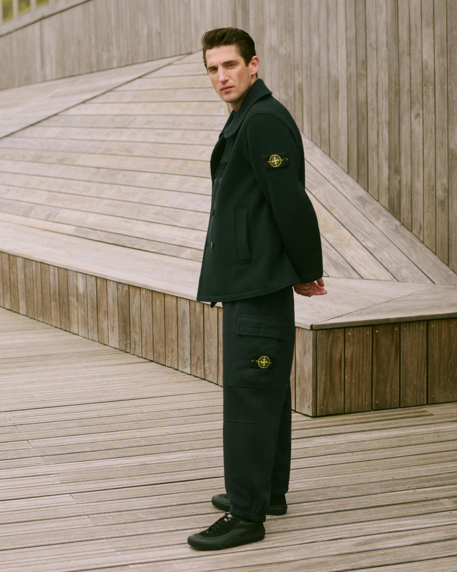 Junger Mann trägt schwarzen Mantel und Hose mit Stone Island Logo, steht vor hellem Holzpodest und Wand.