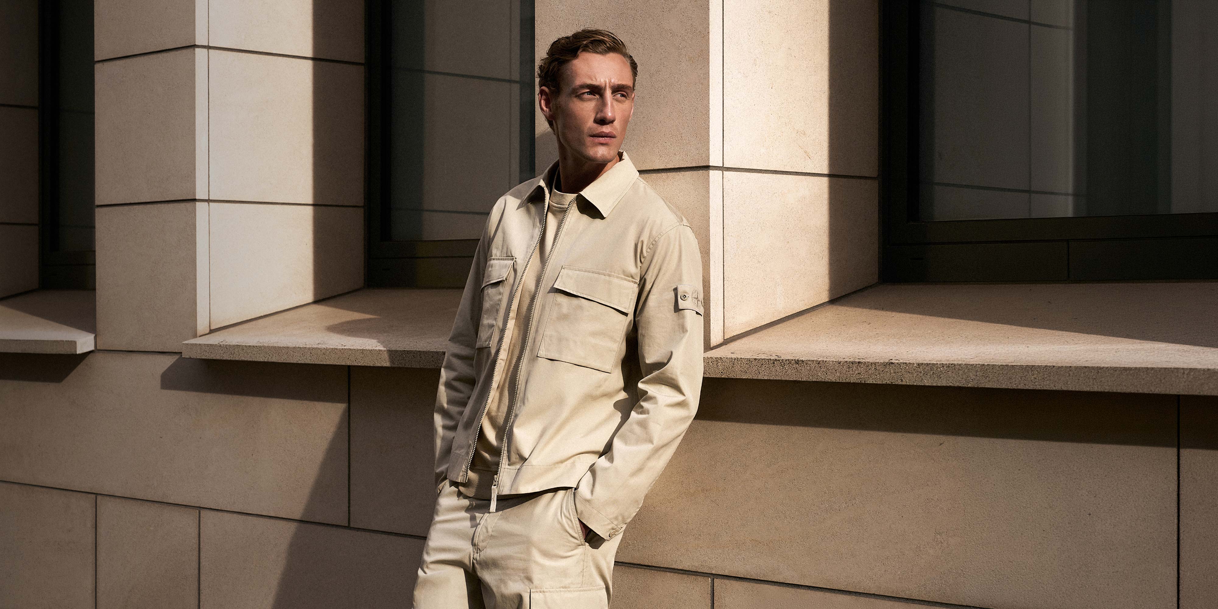 Junger Mann in beige Jacke und Hose lehnt an heller Steinwand mit Schatten, schaut nach rechts, Hände in Taschen.
