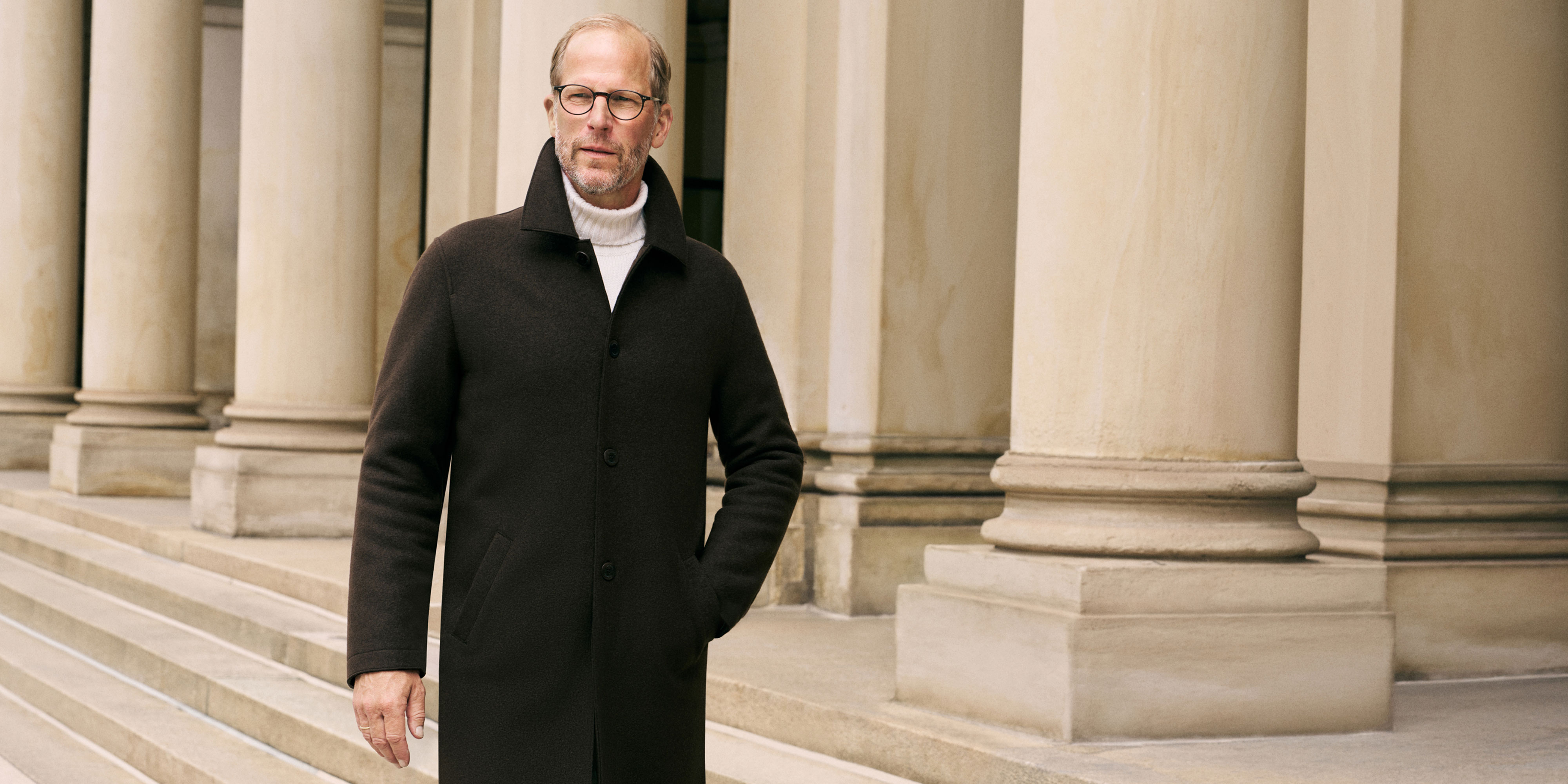Mann mit Brille und Bart trägt dunklen Mantel und weißen Rollkragenpullover, steht vor Säulen eines historischen Gebäudes.