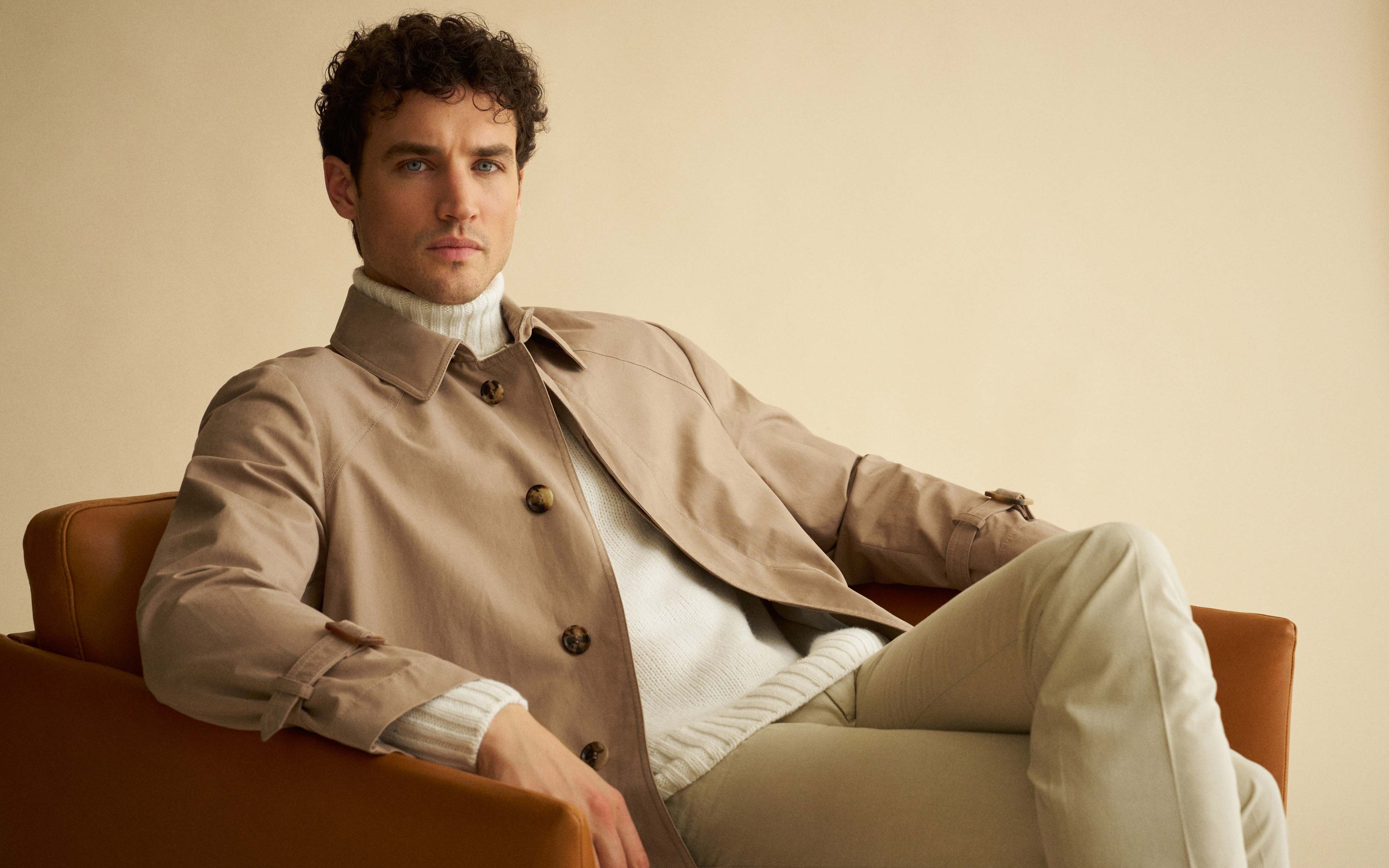 Mann mit lockigem Haar in beiger Jacke und weißem Rollkragenpullover sitzt entspannt auf braunem Sessel vor beige Hintergrund.