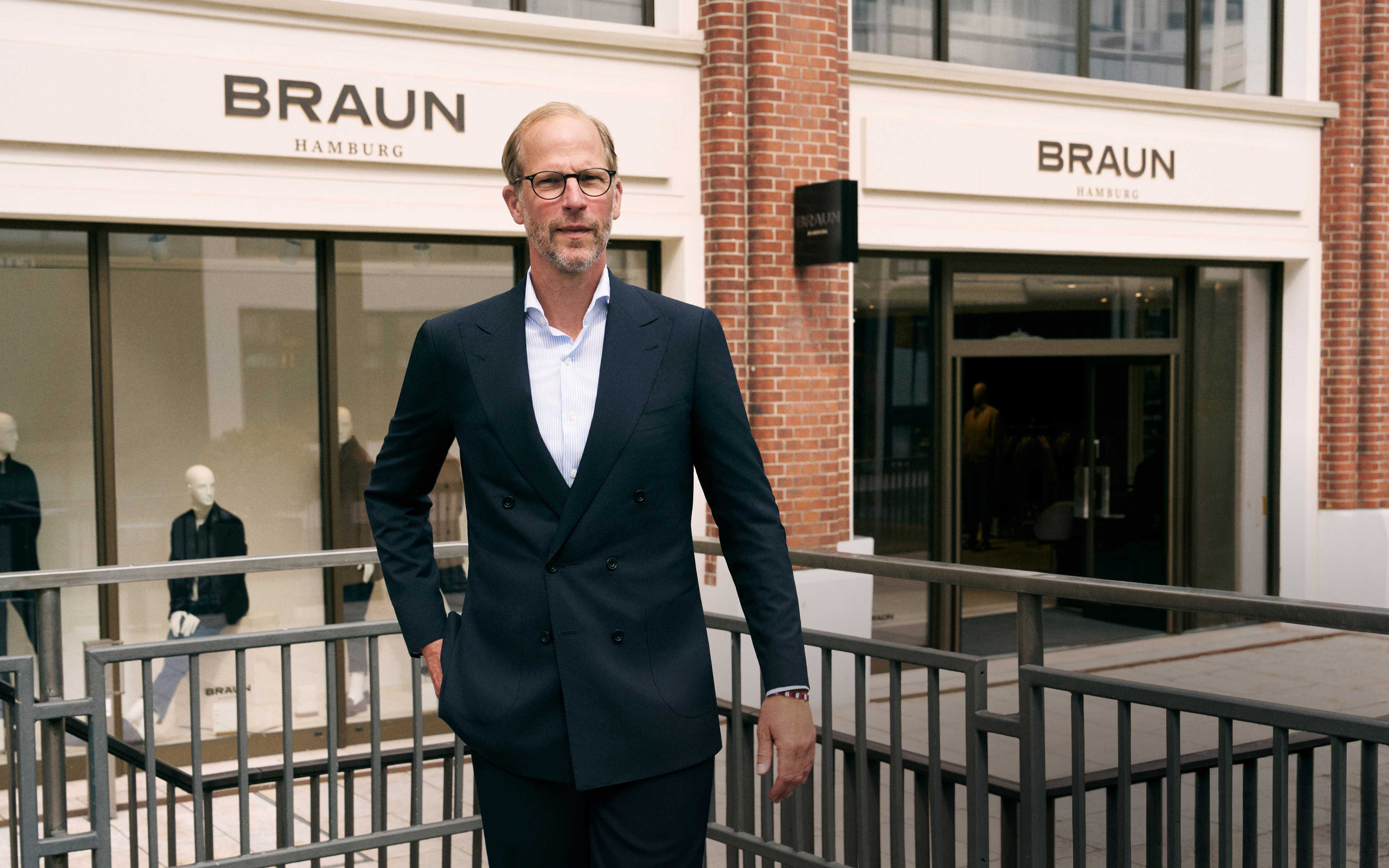 Mann mit Brille und dunkelblauem Anzug steht vor dem Modegeschäft BRAUN Hamburg mit Schaufenster und roter Backsteinfassade.
