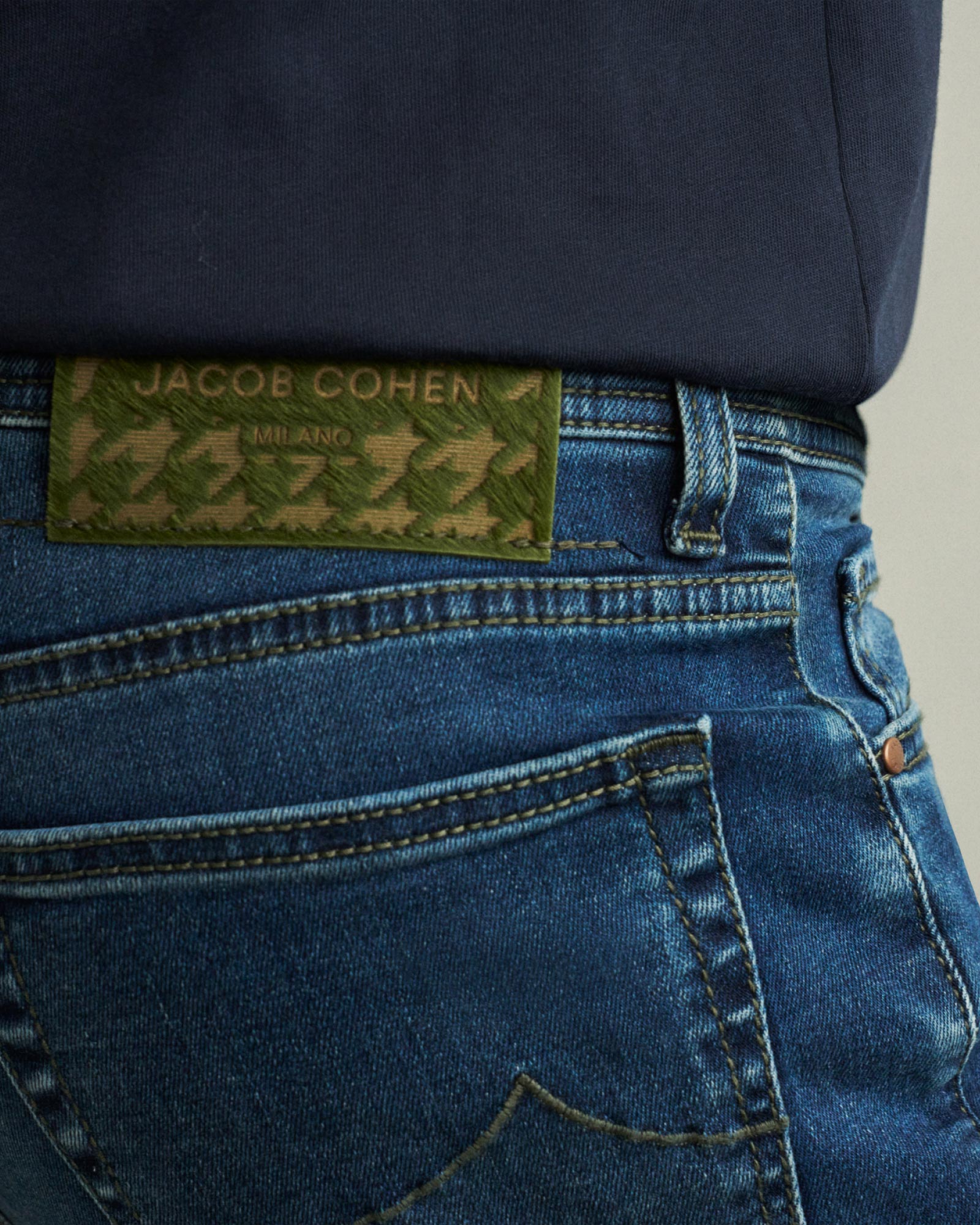 Jeanshose in Blau mit grünem Jacob Cohen Milano Lederetikett und dunkelblauem Oberteil, Detailansicht Bund und Tasche.