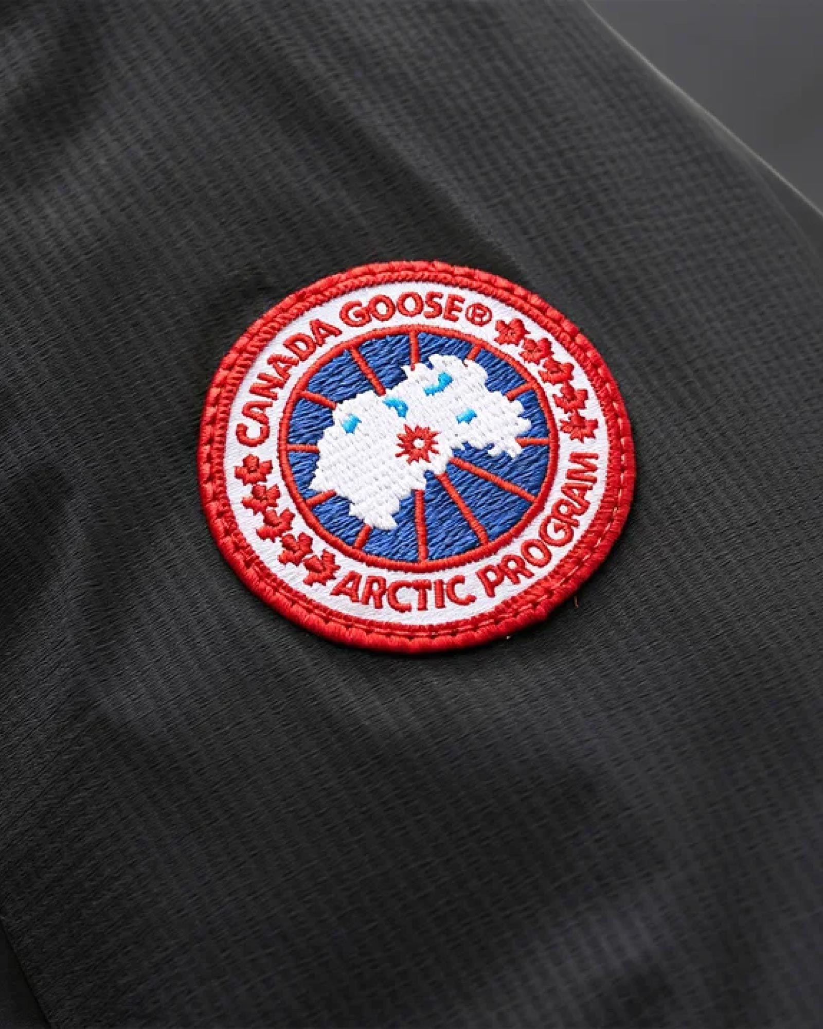 Rundes Canada Goose Logo mit rotem Rand, weißer Schrift und blau-weißem Arktis-Design auf schwarzem Stoff.