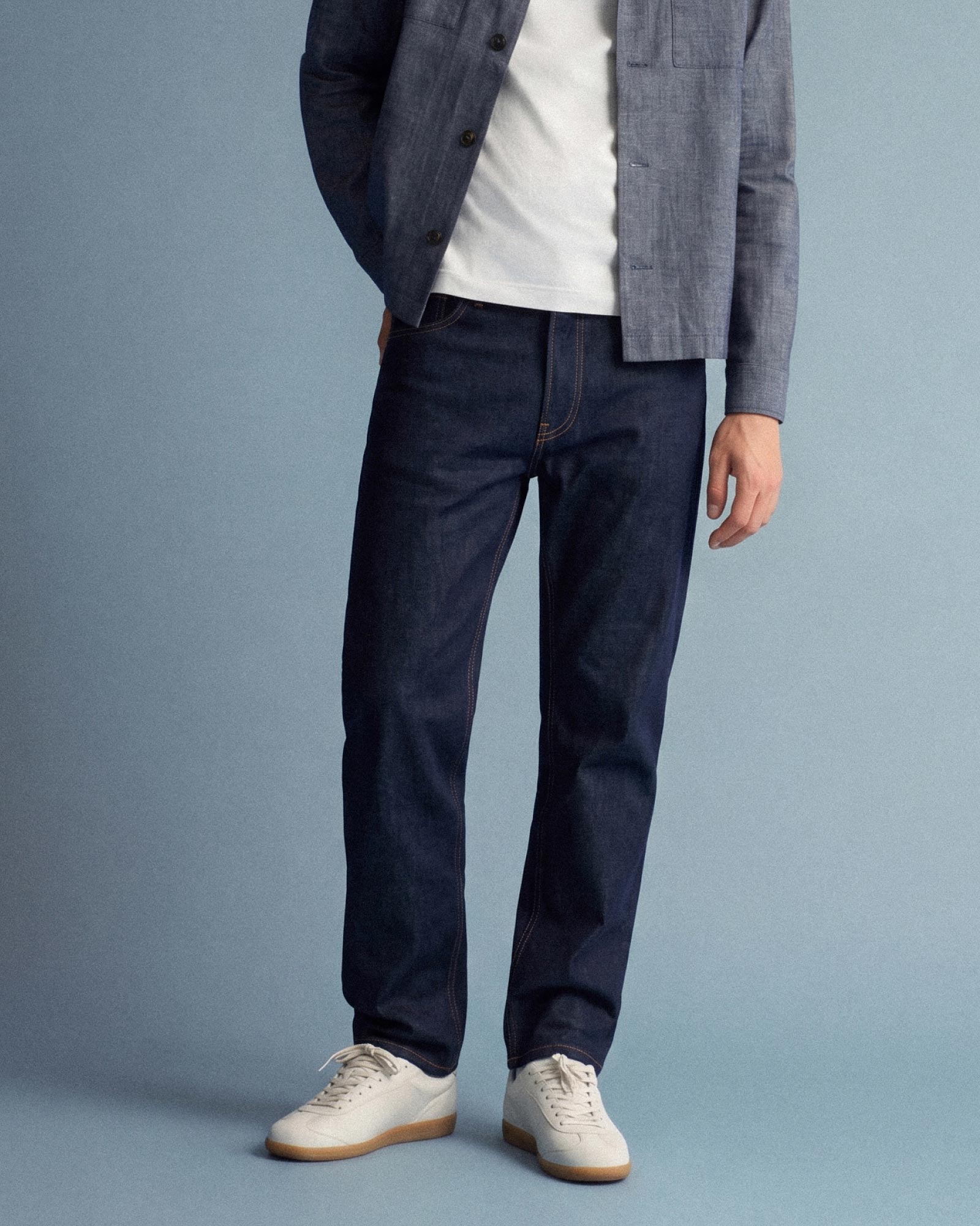 Mann trägt dunkle Jeans, weiße Sneakers, weißes T-Shirt und blaue Hemdjacke vor hellblauem Hintergrund.