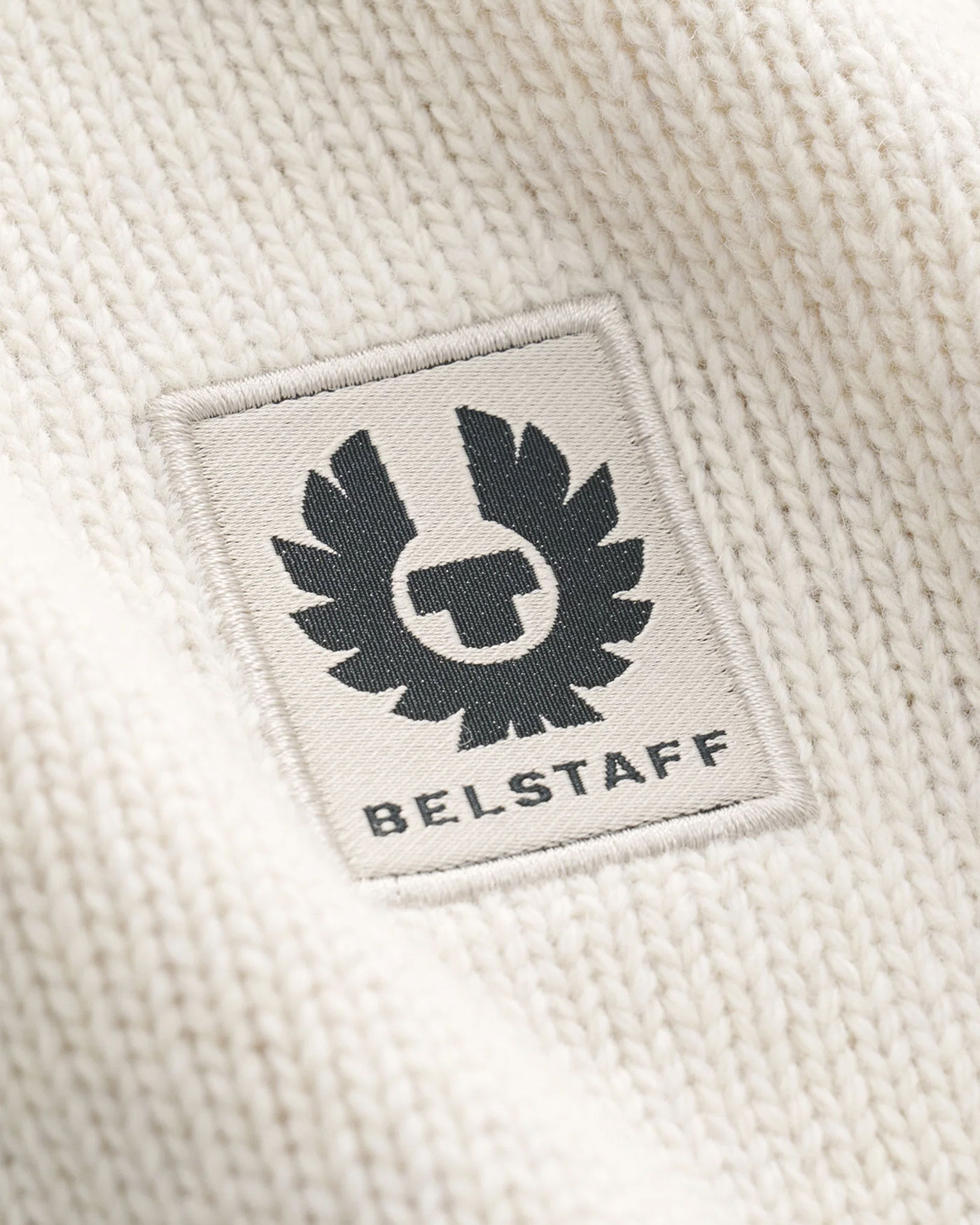 Nahaufnahme eines weißen Strickstoffs mit einem eingenähten rechteckigen Belstaff-Logo in Schwarz und Weiß.