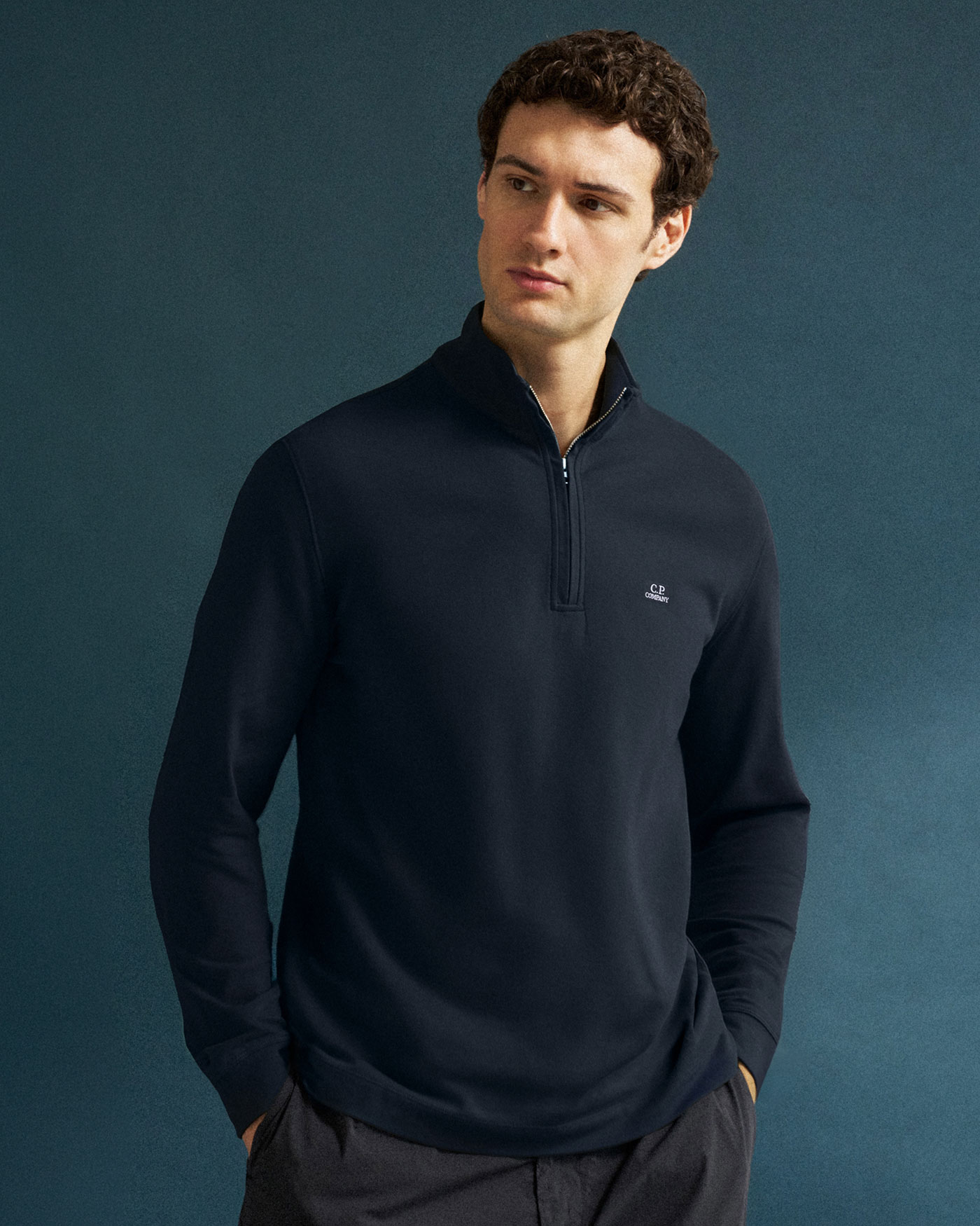 Junger Mann mit dunkelblauem Pullover mit halbem Reißverschluss und C.P. Company Logo vor blauem Hintergrund.