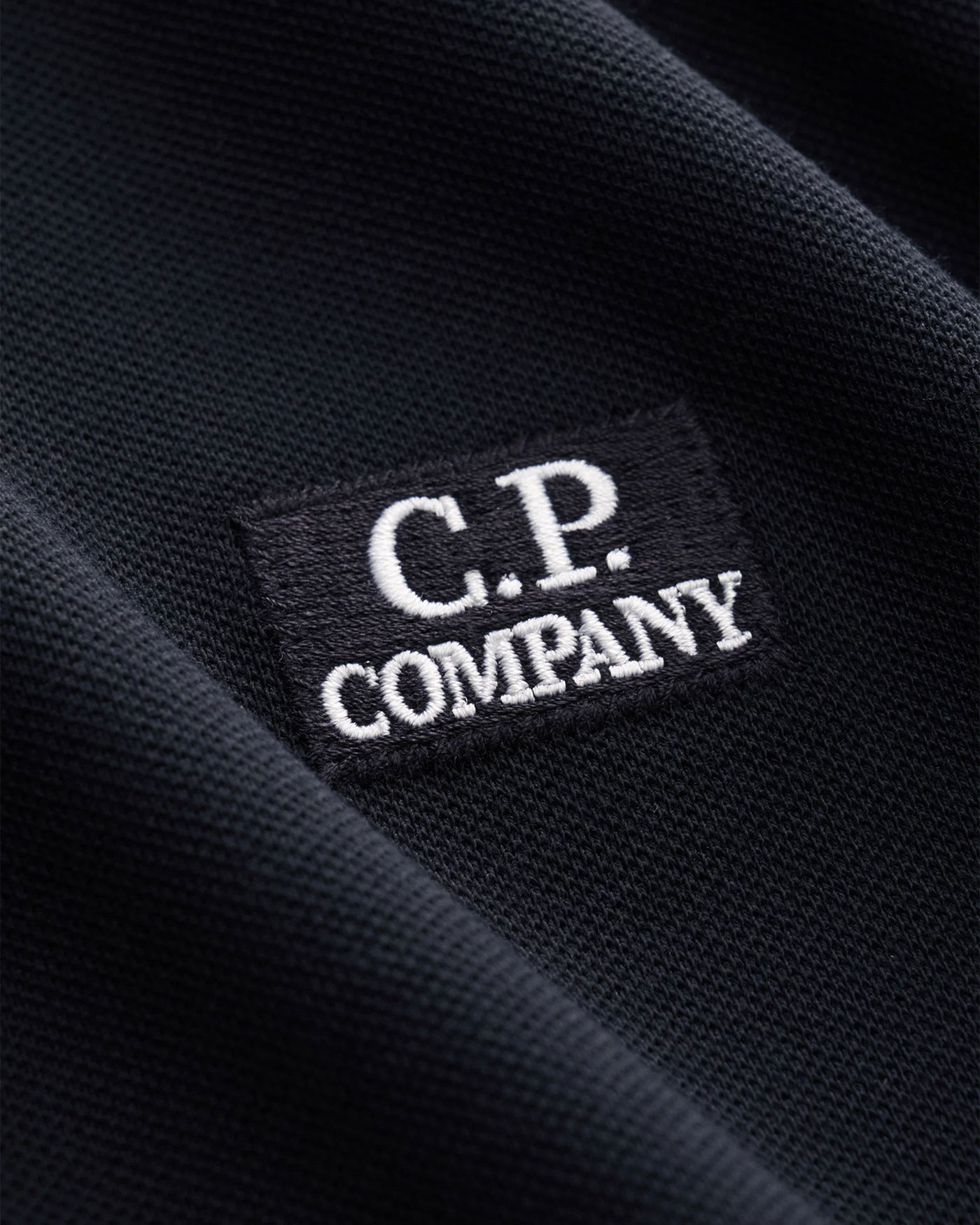 Schwarzes Stoffstück mit eingewebtem schwarzem Label, darauf weißer Schriftzug „C.P. Company“ in Großbuchstaben.