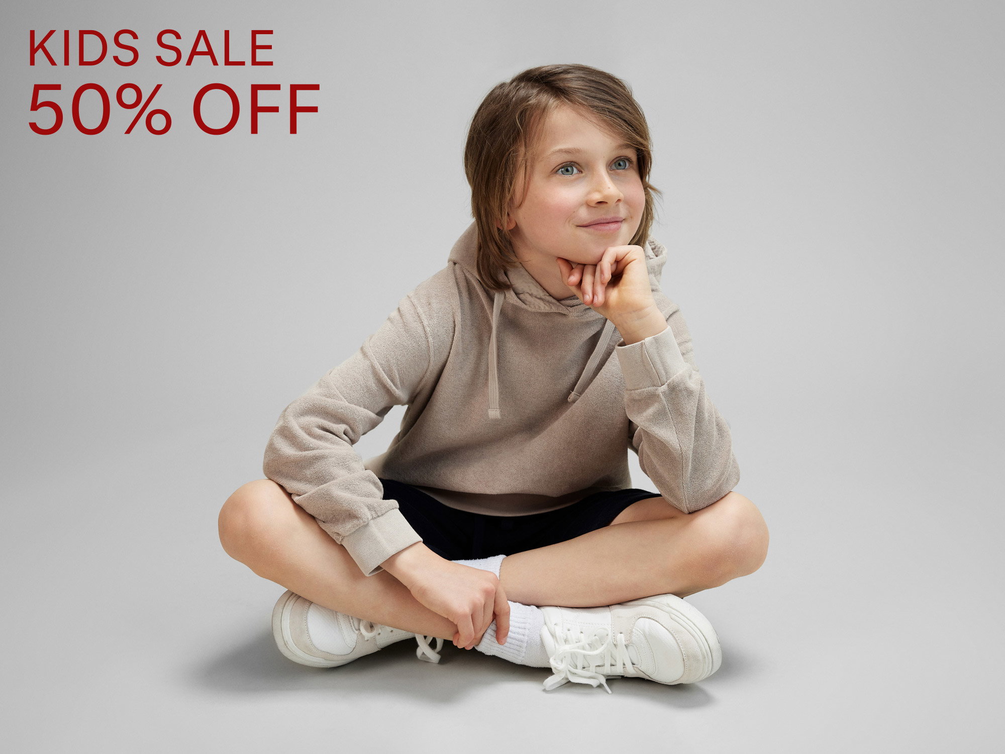 Smiling child in beige hoodie and black shorts sitting cross-legged on gray floor with red text "KIDS SALE 50% OFF" on left side.