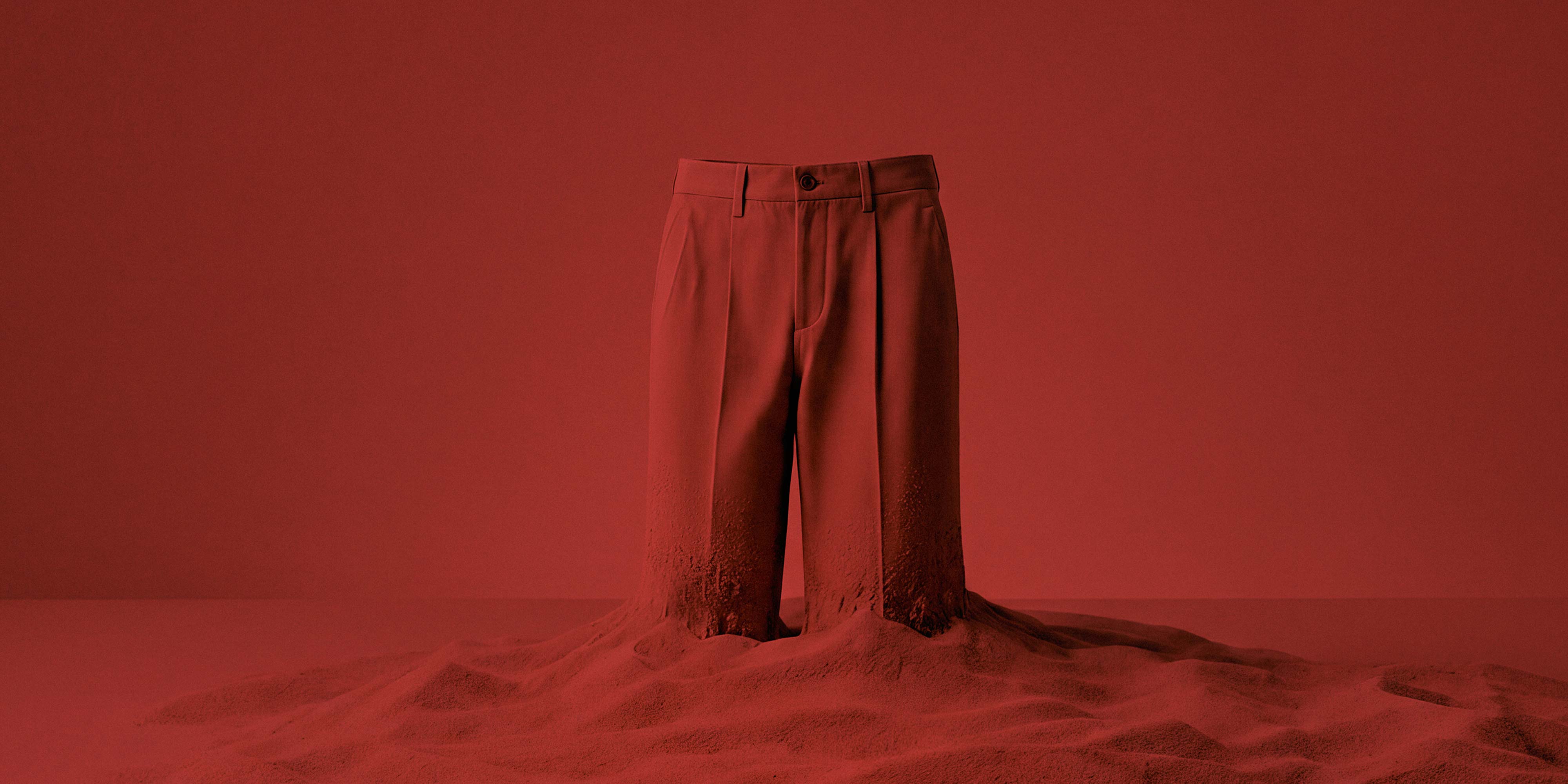 Rote Anzughose steht auf rotem Sand, der um die Hosenbeine herum auftürmt, minimalistisches monochromes Setting.