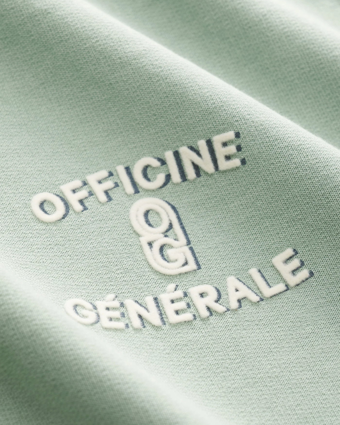 Nahaufnahme eines hellgrünen Baumwollstoffs mit weißer und dunkelblauer Stickerei "Officine Générale" und Logo.