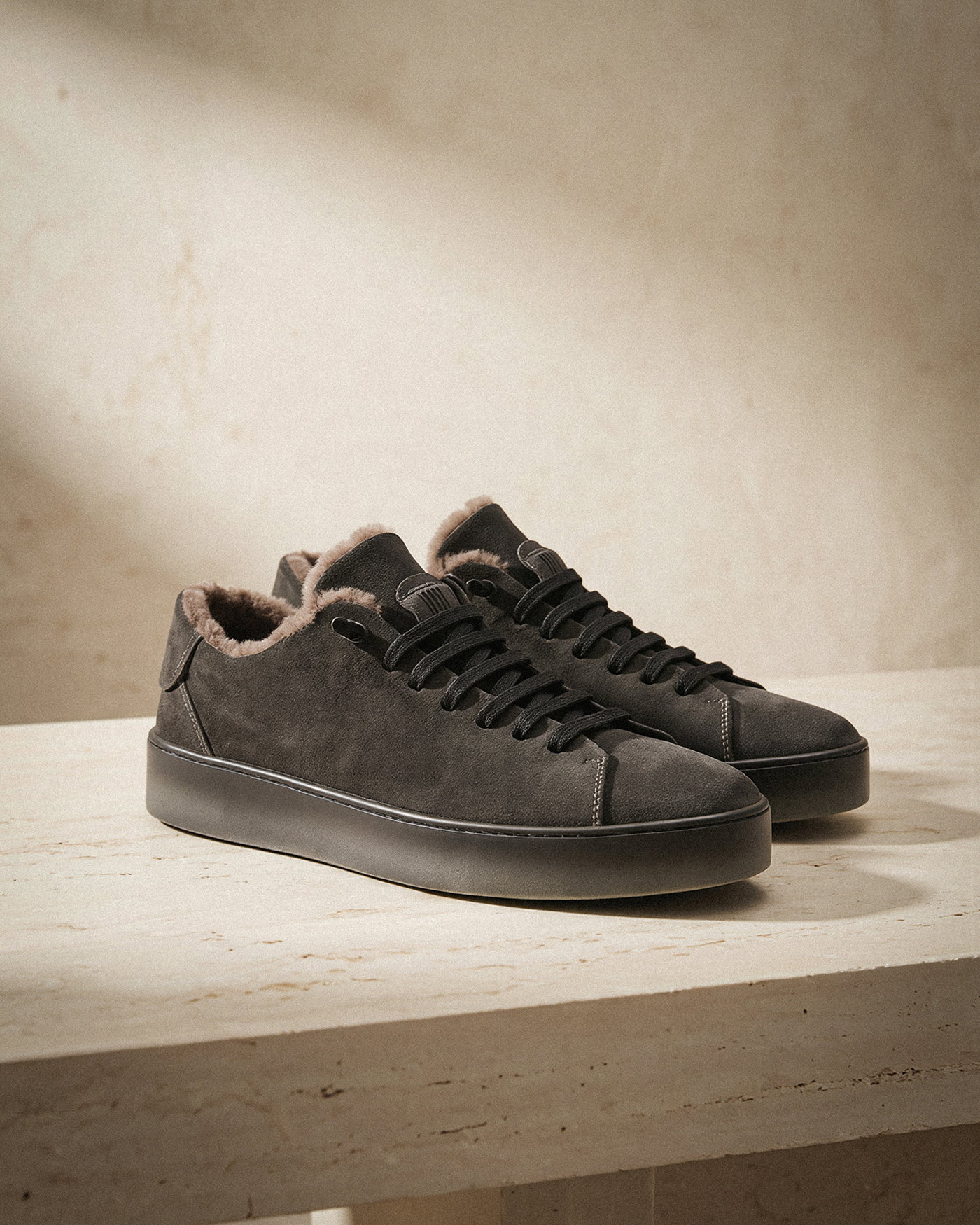Schwarze Sneaker aus Wildleder mit dicker Sohle und warmem, hellbraunem Fellfutter stehen auf einem hellen Marmortisch.