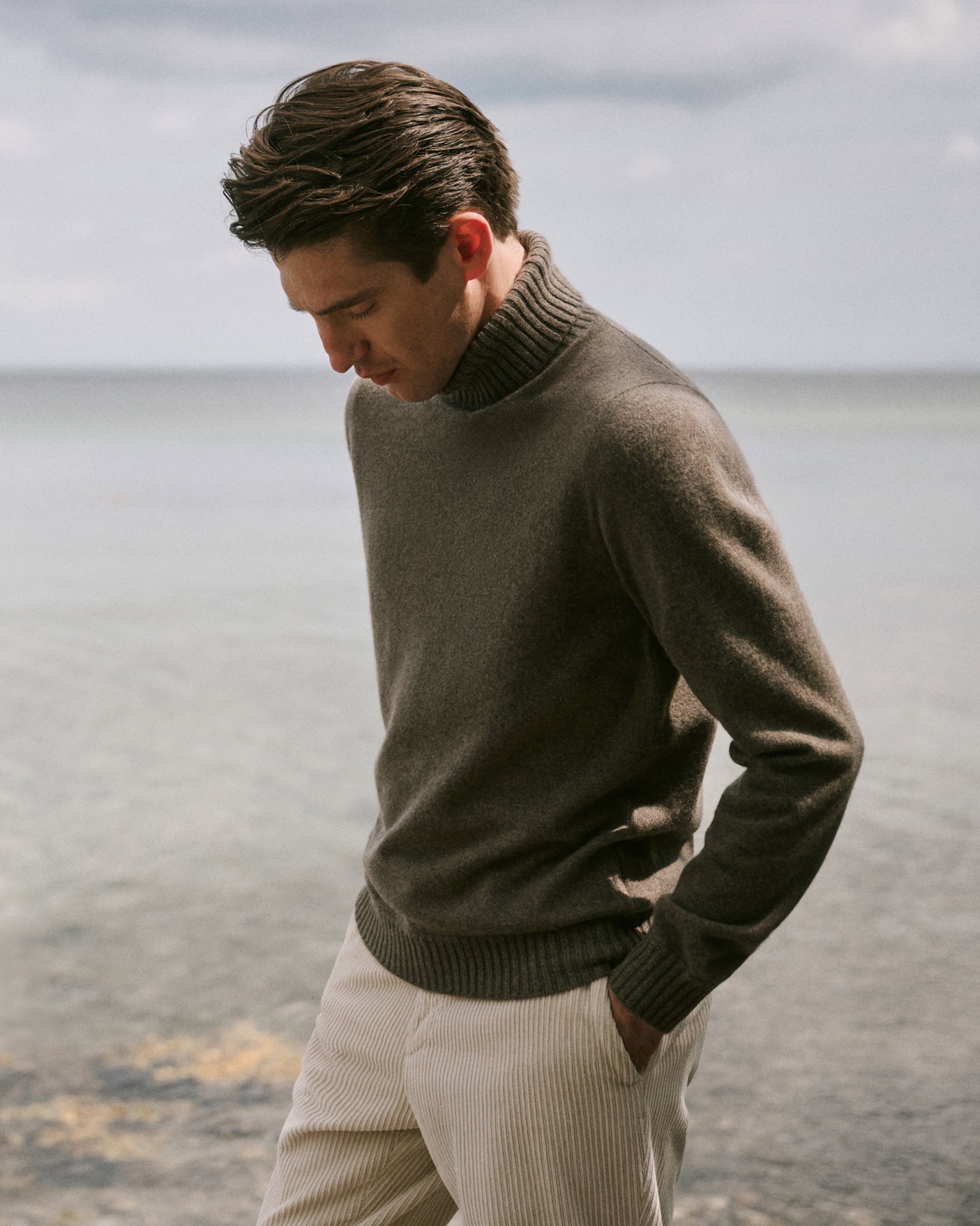 Mann mit dunkelbraunem Rollkragenpullover und hellen Cordhosen steht am Wasser, Blick nach unten, Hände in den Taschen.