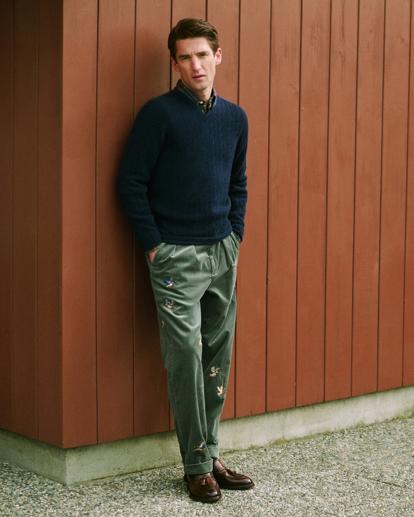 Mann lehnt an roter Holzfassade, trägt dunkelblauen V-Neck Pullover, kariertes Hemd, grüne Kordhose mit Vogelstickerei, braune Schuhe.