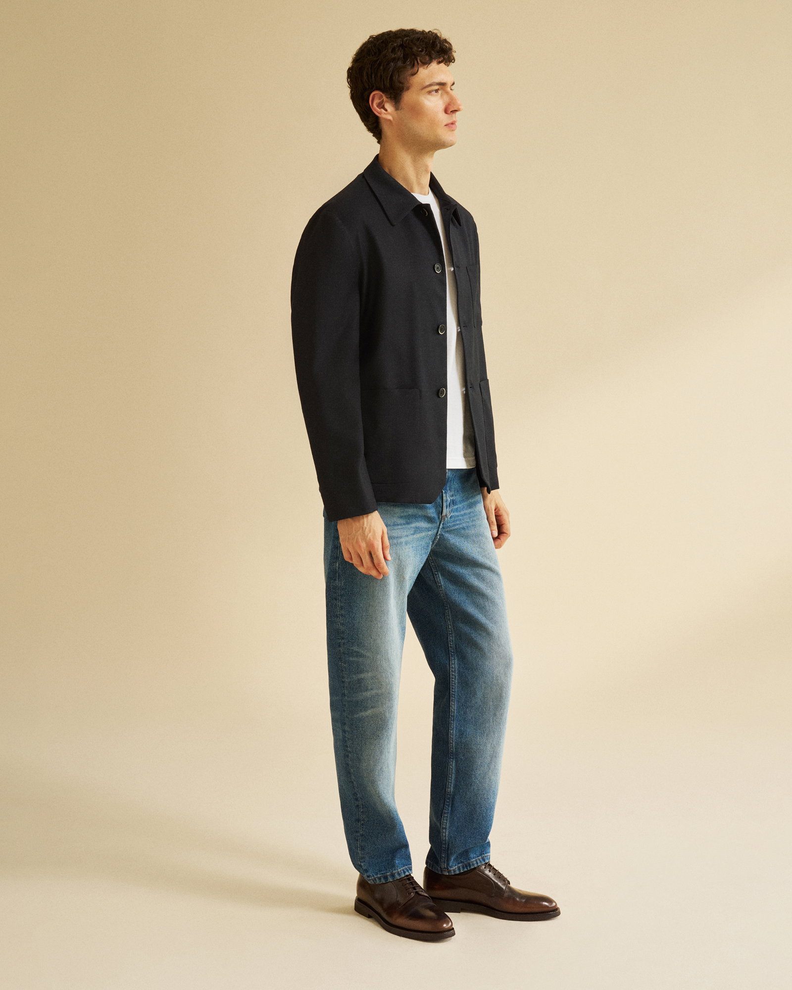 Mann trägt schwarze Jacke, weißes T-Shirt, blaue Jeans und braune elegante Anzugschuhe, steht vor beigem Hintergrund.