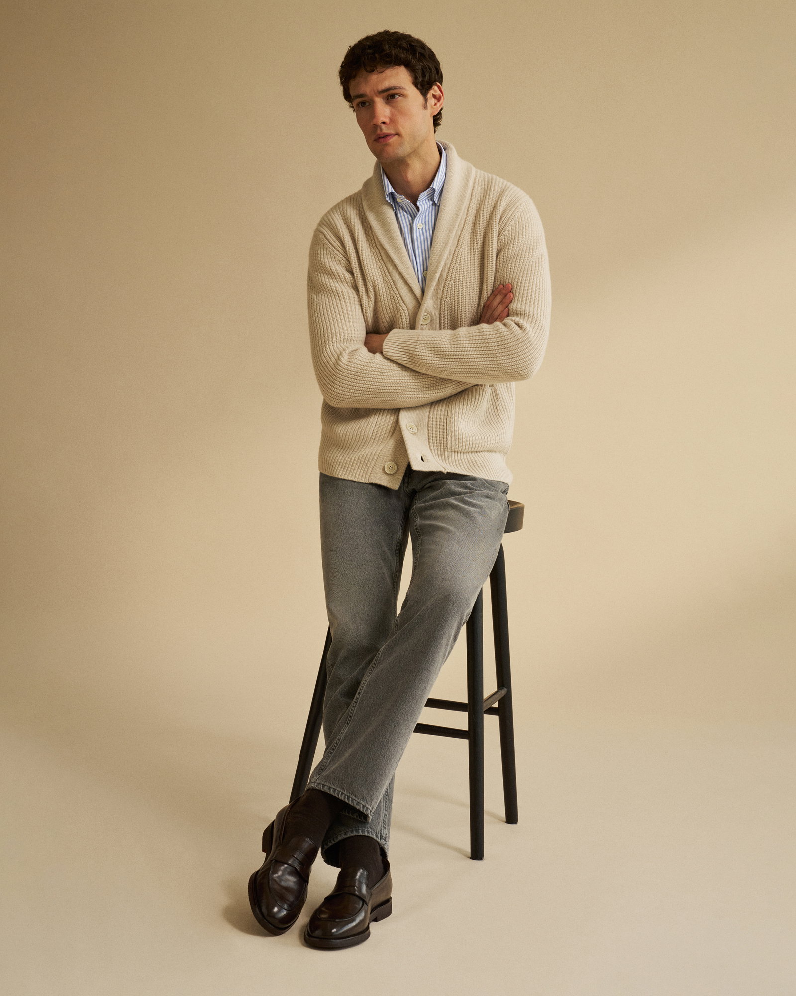 Junger Mann sitzt auf schwarzem Hocker, trägt beige Strickjacke, blau-weiß gestreiftes Hemd, graue Jeans und braune Loafer Schuhe.
