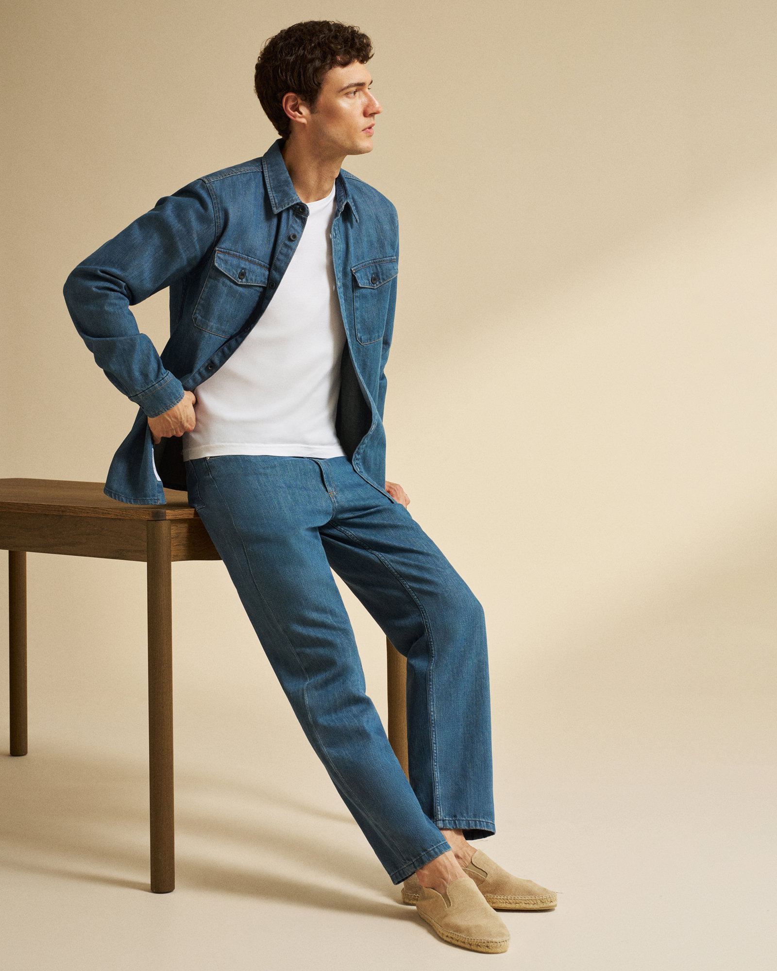 Junger Mann sitzt auf Holztisch, trägt Jeansjacke, weiße T-Shirt, blaue Jeans und beige Slipper, blickt seitlich.