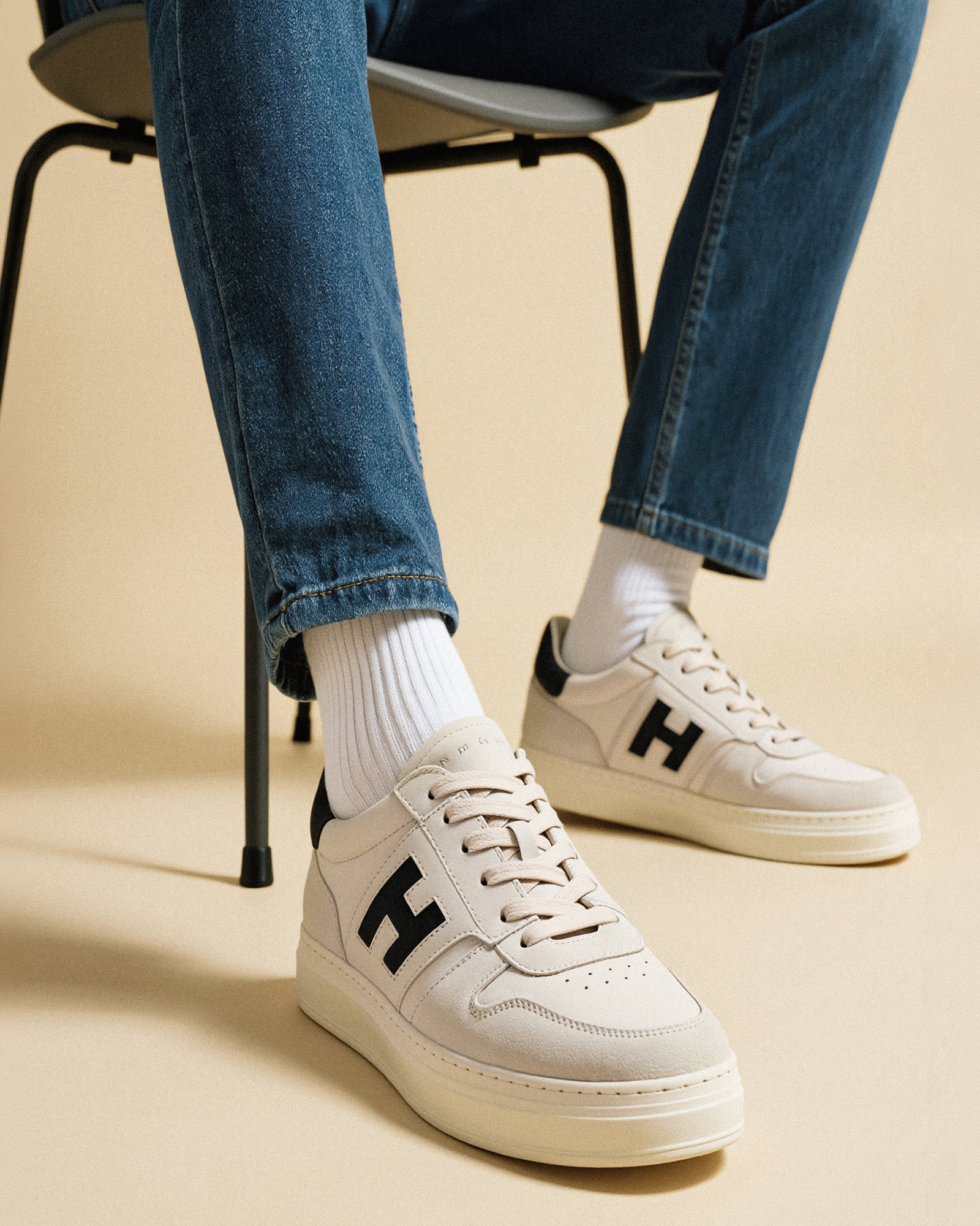 Weißer Sneaker mit schwarzem "H"-Logo, weißer Socken und blauen Jeans, sitzend auf schwarzem Stuhl vor beigem Hintergrund.