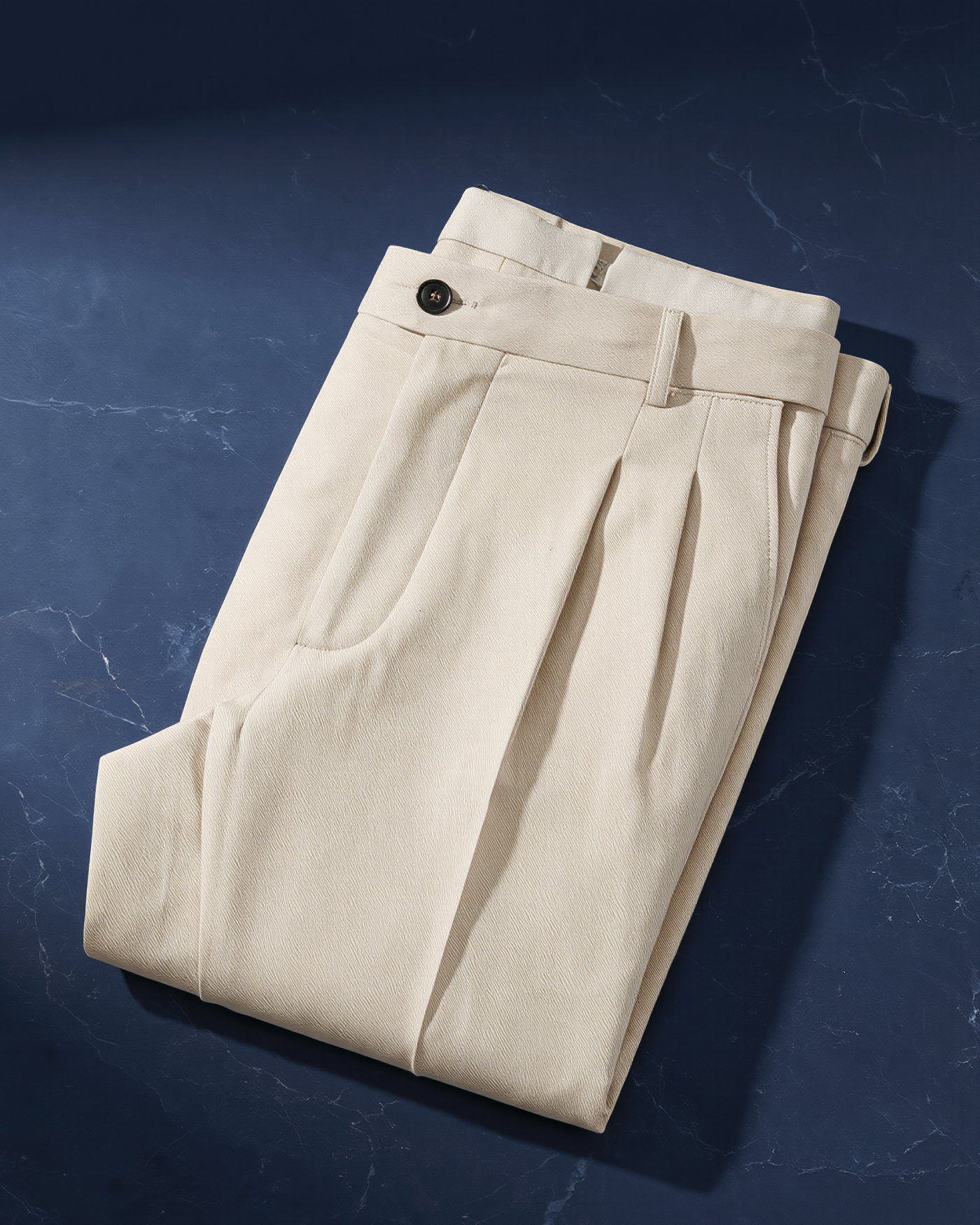 Beige Chino-Hose mit doppelter Bundfalte, Knopfverschluss und Gürtelschlaufen, ordentlich gefaltet auf dunkelblauem Untergrund.