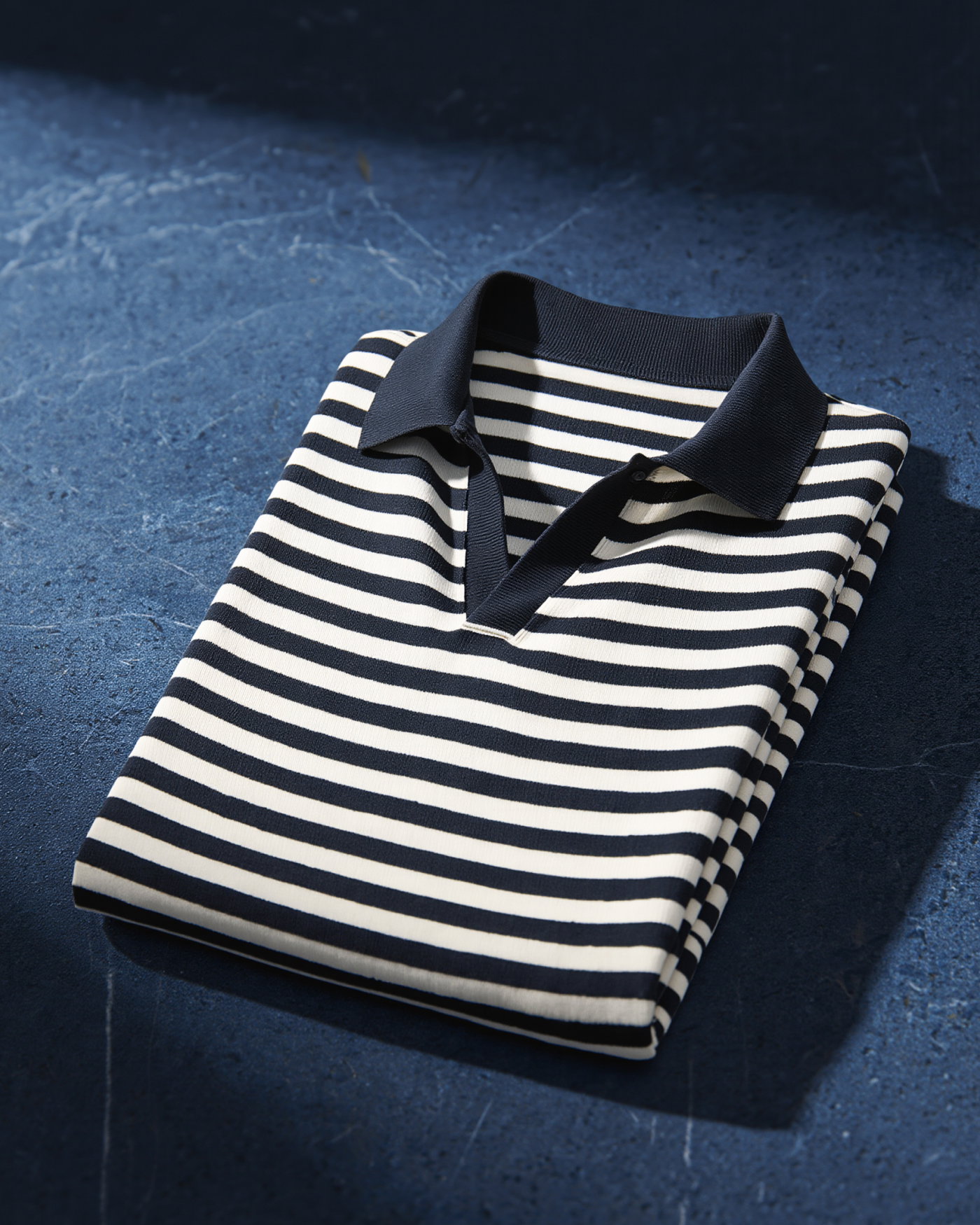 Gestapeltes Polo-Langarmshirt mit navy-weiß horizontalen Streifen und offenem, dunkelblauem Kragen auf blauem Untergrund.