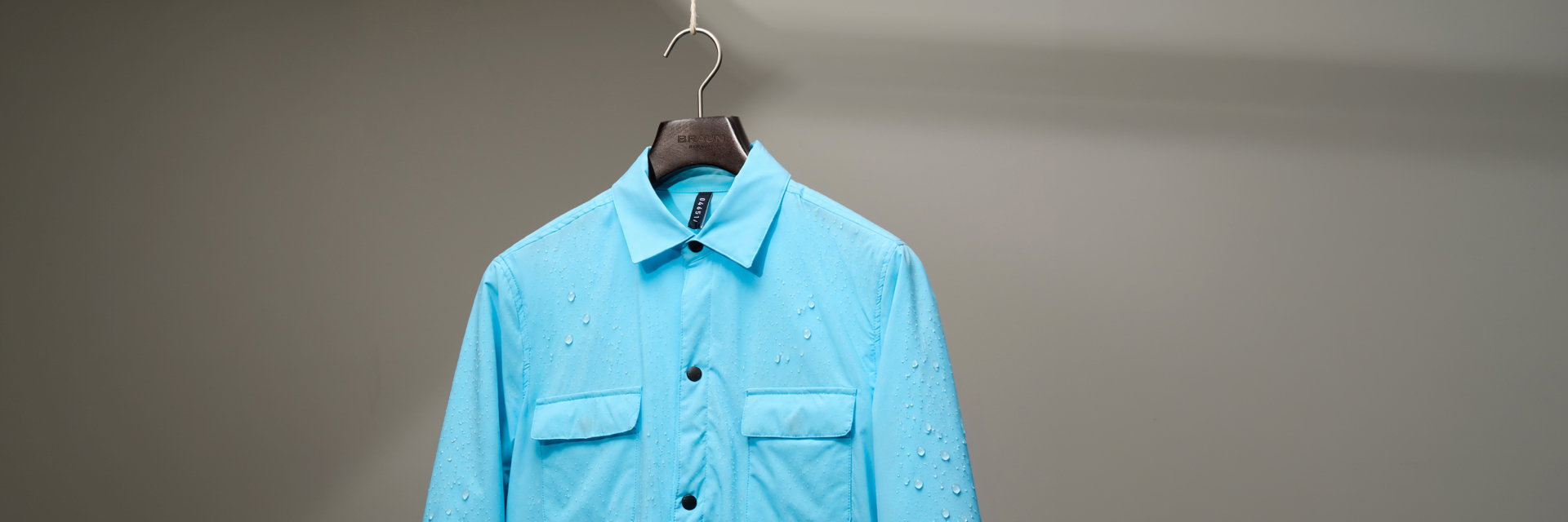 Hellblaue Herren-Regenjacke mit Kragen und Brusttaschen, hängend an einem Kleiderbügel, mit abperlenden Regentropfen.