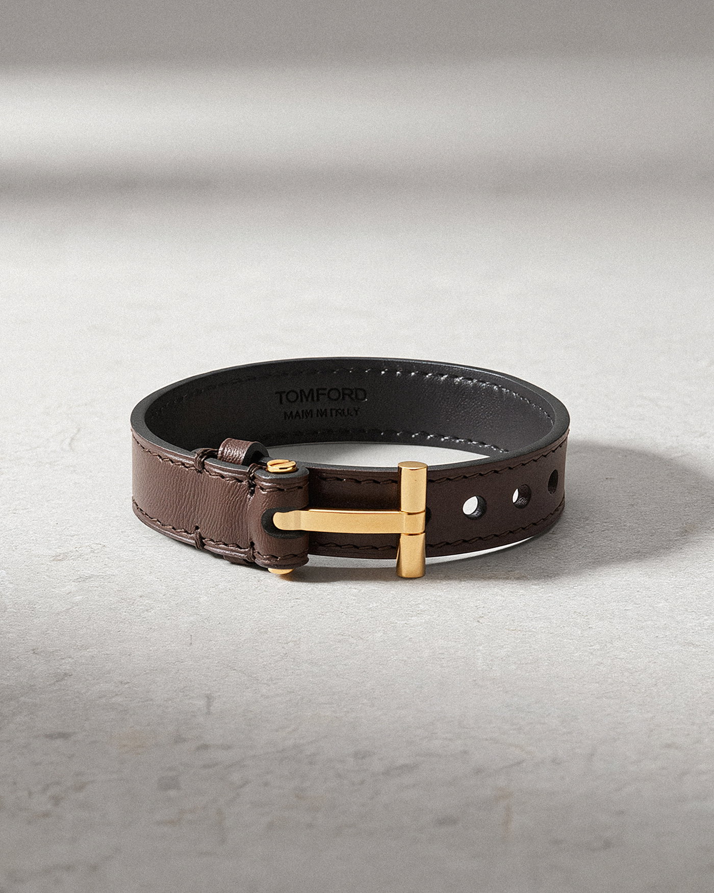 Braunes Herren Lederarmband mit goldfarbenem Verschluss und Lochung, liegend auf hellem Untergrund, Innenseite mit TOM FORD Schriftzug.