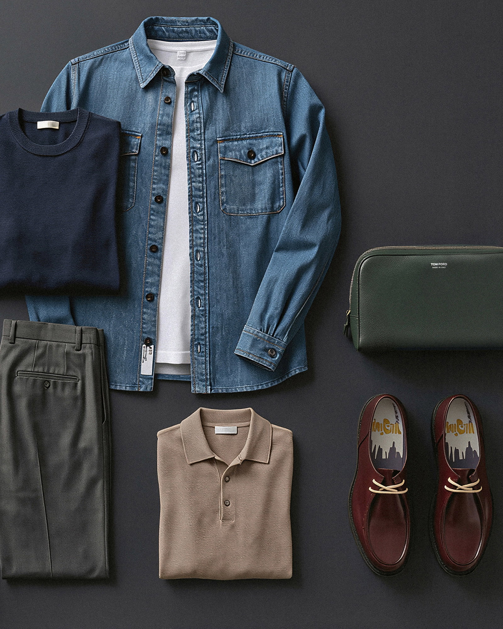 Auf dem Boden liegendes Herrenoutfit mit Jeanshemd, weißem T-Shirt, navy Pullover, grauer Hose, beige Polo, braunen Schuhen und grüner Tasche.