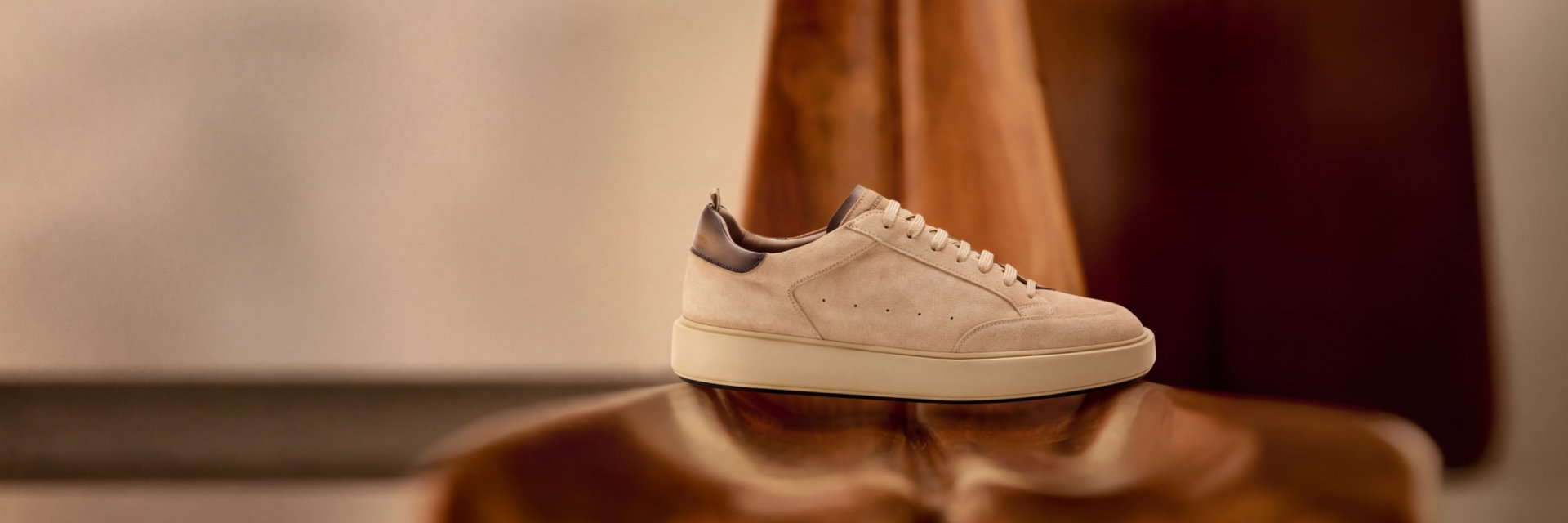 Beiger Herrensneaker aus Veloursleder mit braunen Akzenten, weißer Sohle, auf glänzendem Holzstuhl präsentiert.