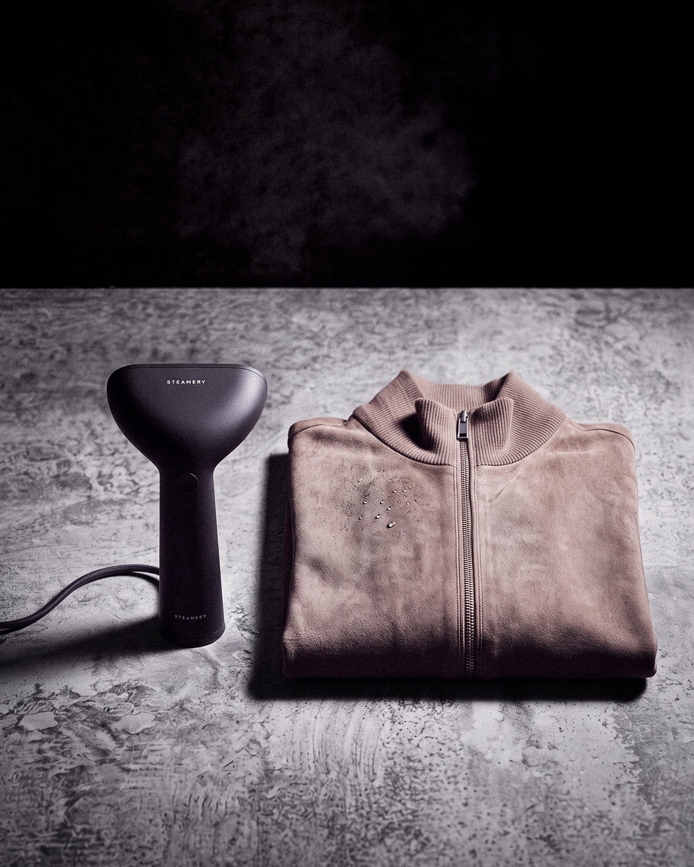 Gefaltete beige Herren-Wildlederjacke mit Reißverschluss auf grauem Tisch neben anthrazitfarbenem Hand-Steamer.