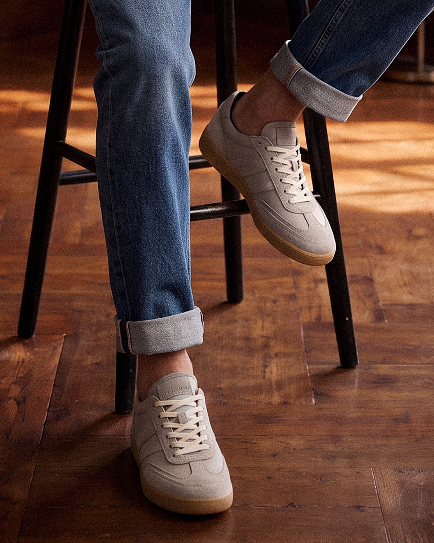 Beige Wildledersneaker mit weißer Sohle zu hellblauen Herrenjeans, Hosenbeine gekrempelt, sitzend auf schwarzem Stuhl.