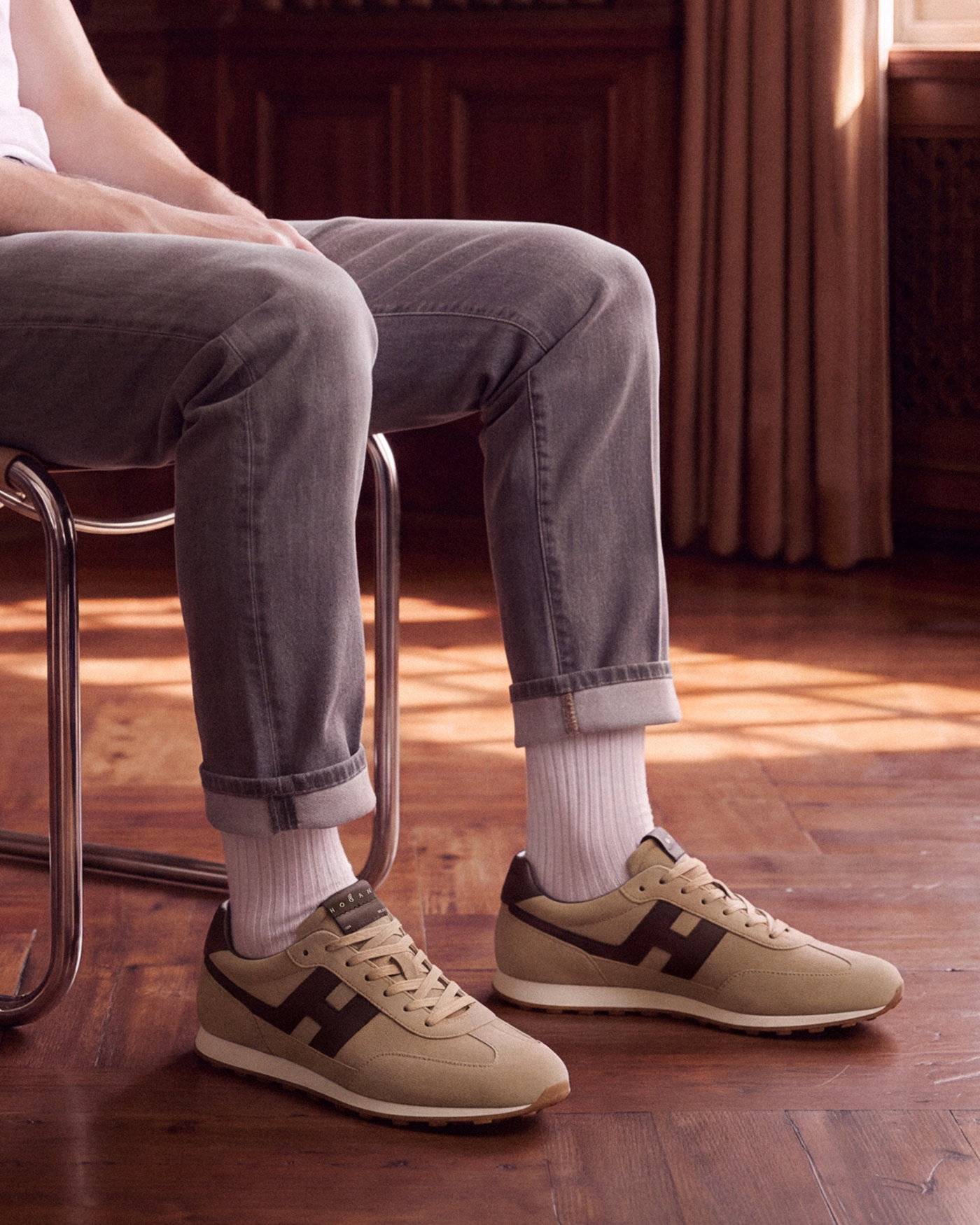 Beine eines sitzenden Mannes mit hellgrauer Jeans, weißen Socken und beige-braunen Wildledersneakern auf Holzfußboden.