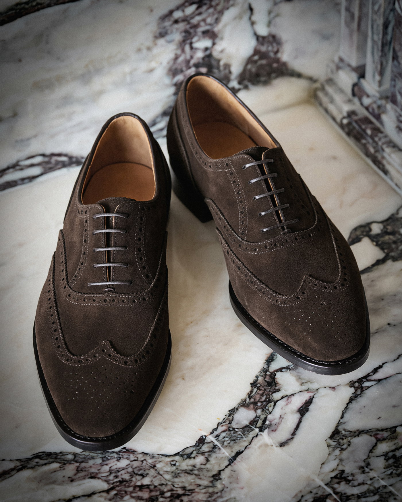 Braune Veloursleder-Herrenschnürschuhe mit Brogue-Details auf weiß-schwarzem Marmorboden fotografiert.
