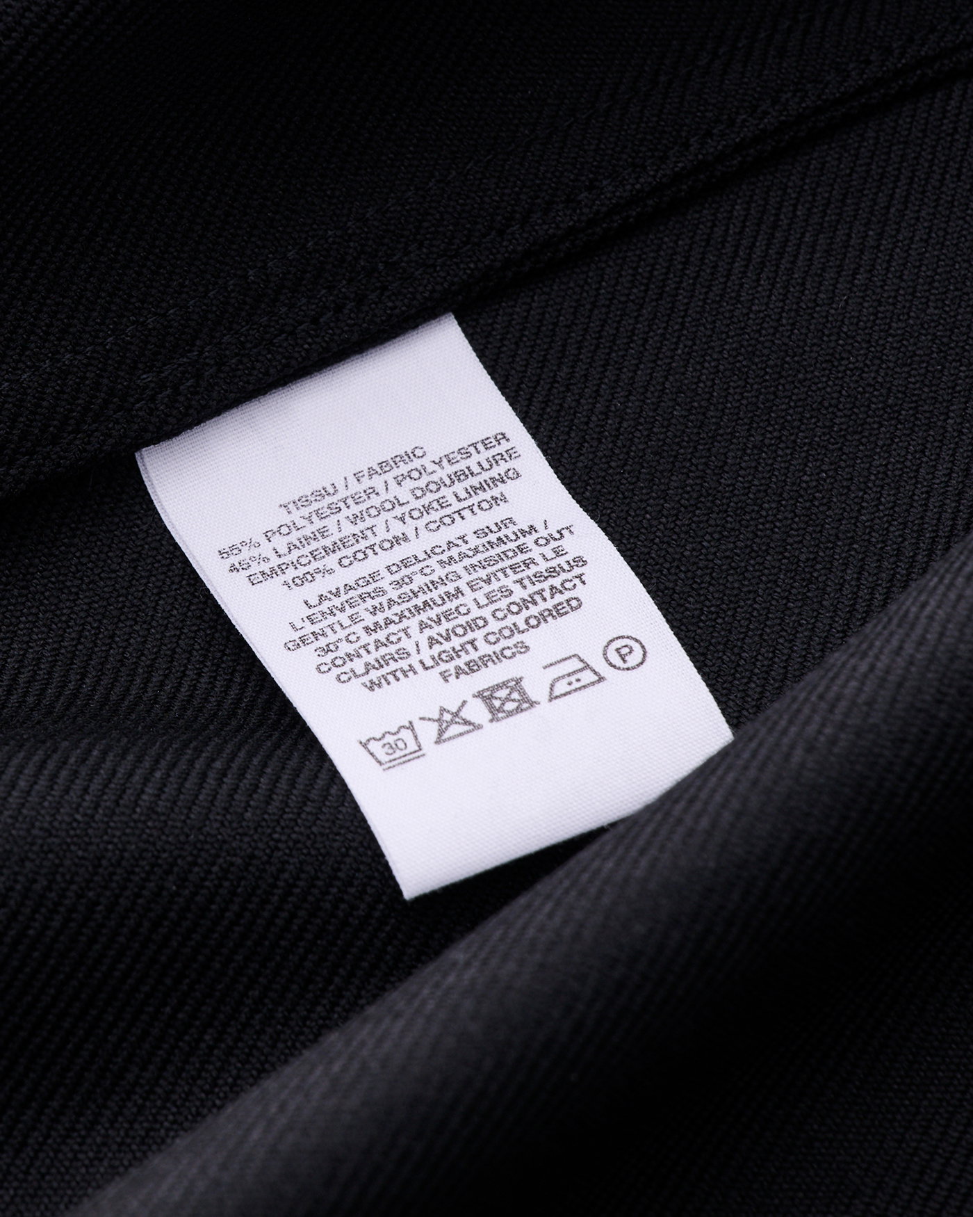 Detailaufnahme eines weißen Waschetiketts mit Pflegehinweisen auf dunkelblauem Polyesterstoff eines Kleidungsstücks.