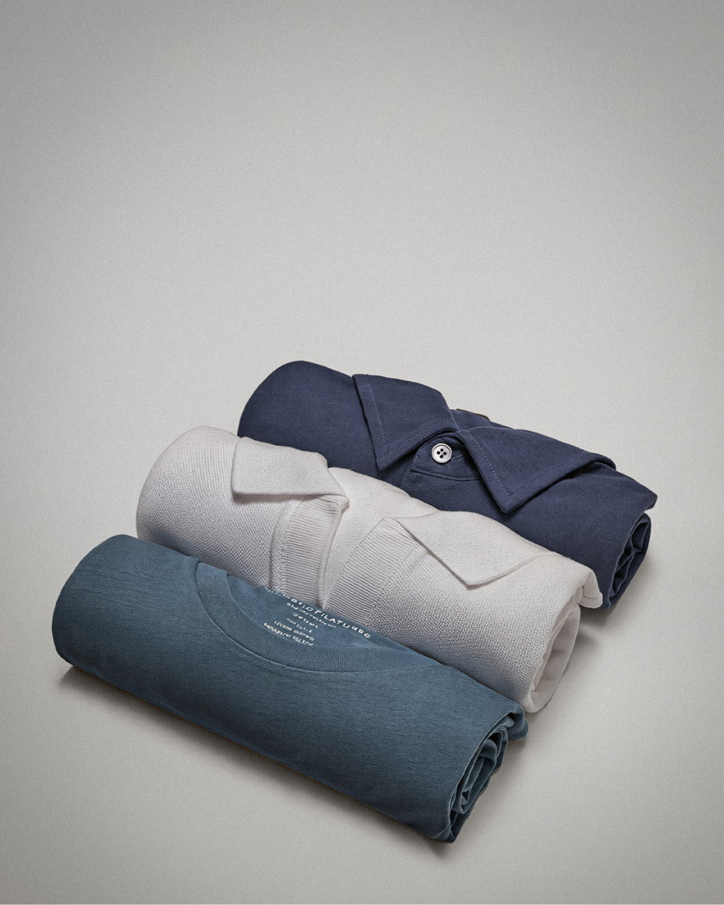 Drei gerollte Herren-Shirts in Dunkelblau mit Kragen, Weiß mit Kragen und Petrol ohne Kragen auf hellem Untergrund.