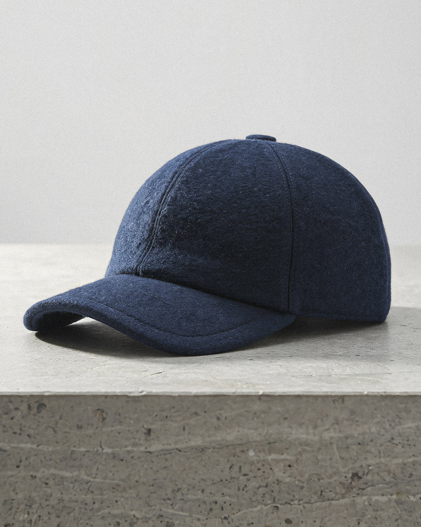 Dunkelblaue Herren-Cap aus weichem Stoff liegt auf grauem, rauem Steinuntergrund, Vorderansicht mit Schirm sichtbar.