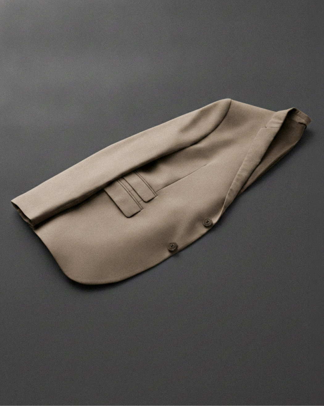 Beiges, in der Mitte gefaltetes Herren-Sakko mit seitlichen Taschen und zwei sichtbaren Knöpfen, auf dunklem Untergrund liegend.