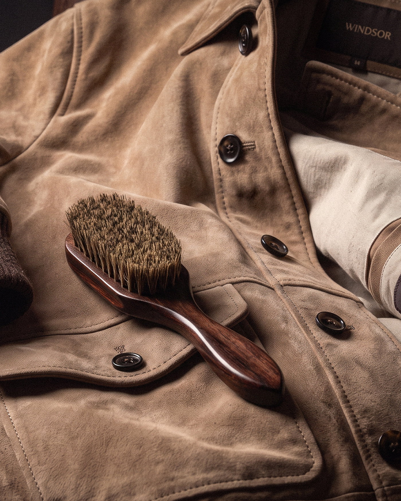 Braune Wildlederbürste mit Holzgriff liegt auf brauner Velourslederjacke mit schwarzen Knöpfen und Innenfutter.