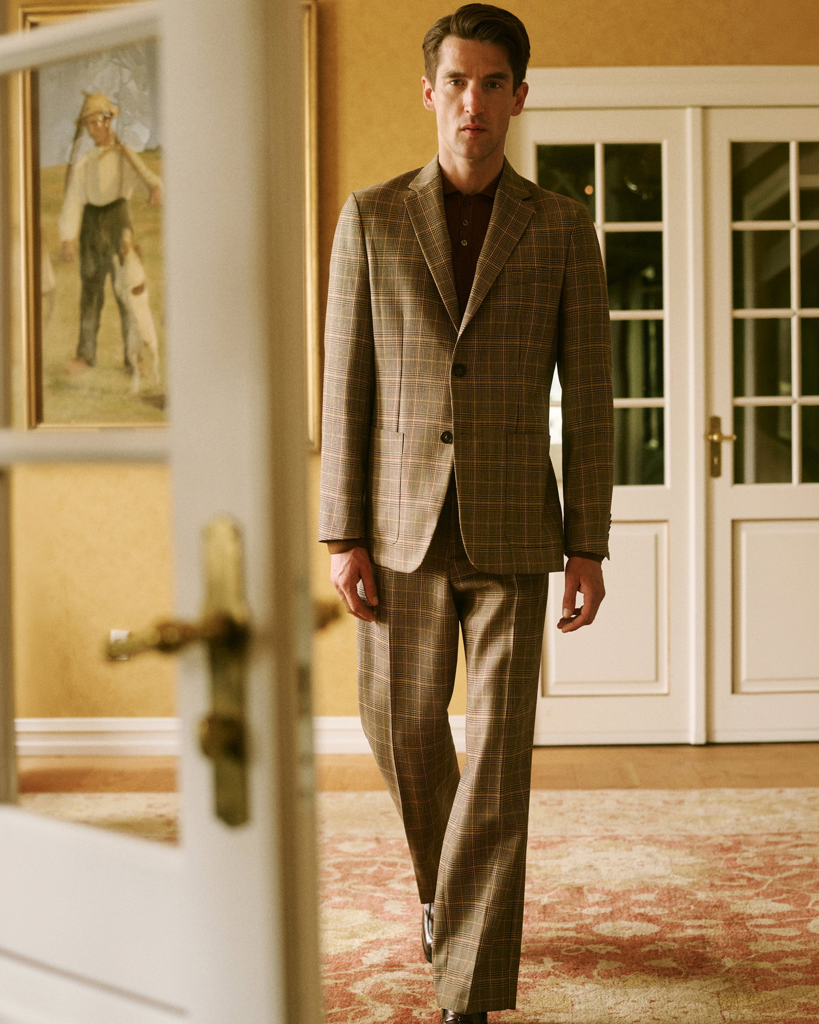 Eleganter Mann in braun-beige kariertem Anzug und dunkelrotem Strickpolo geht durch ein klassisch eingerichtetes Zimmer.