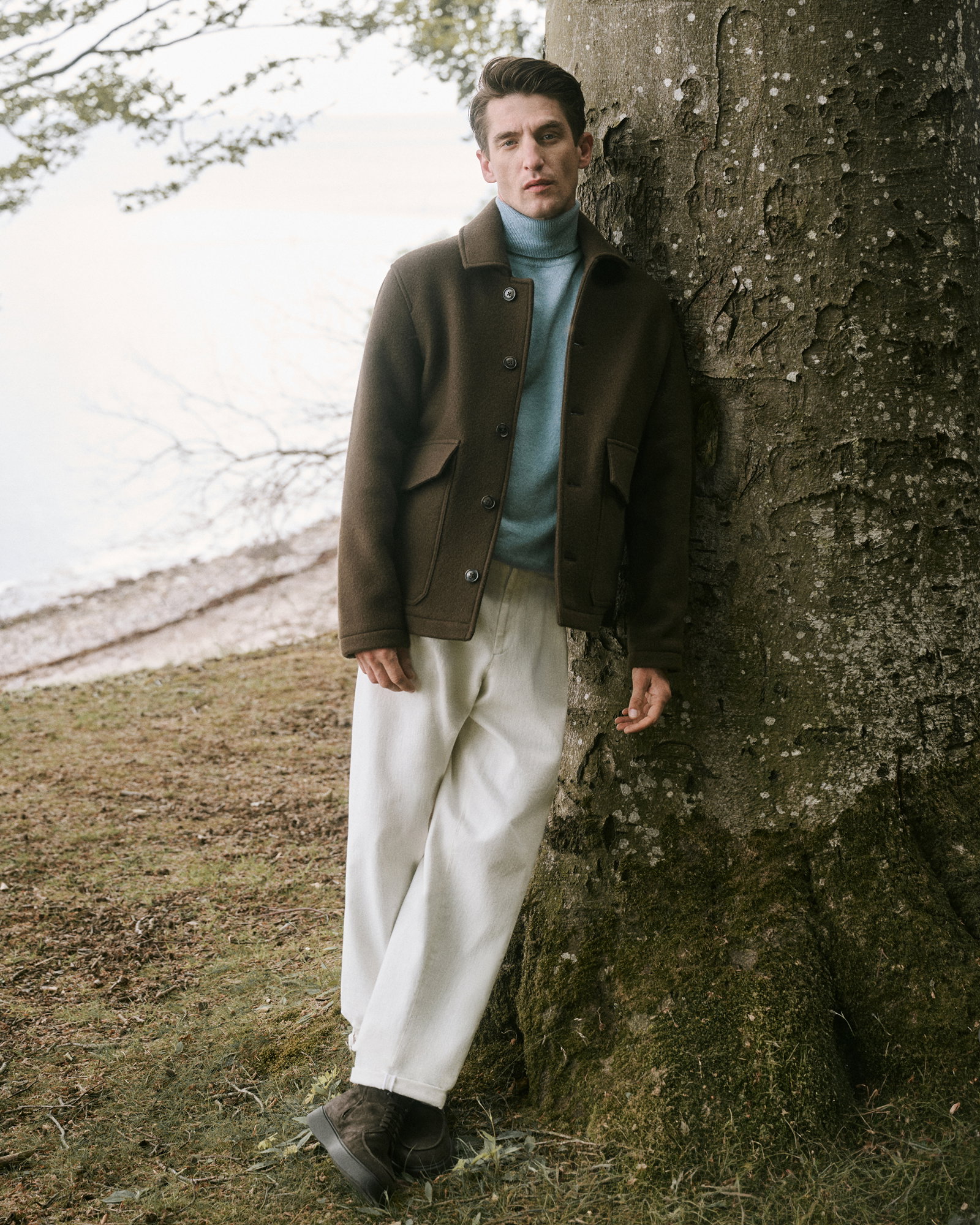 Mann lehnt an Baum, trägt braune Jacke, hellblauen Rollkragenpullover, beige Hose und braune Schuhe, Wald und See im Hintergrund.