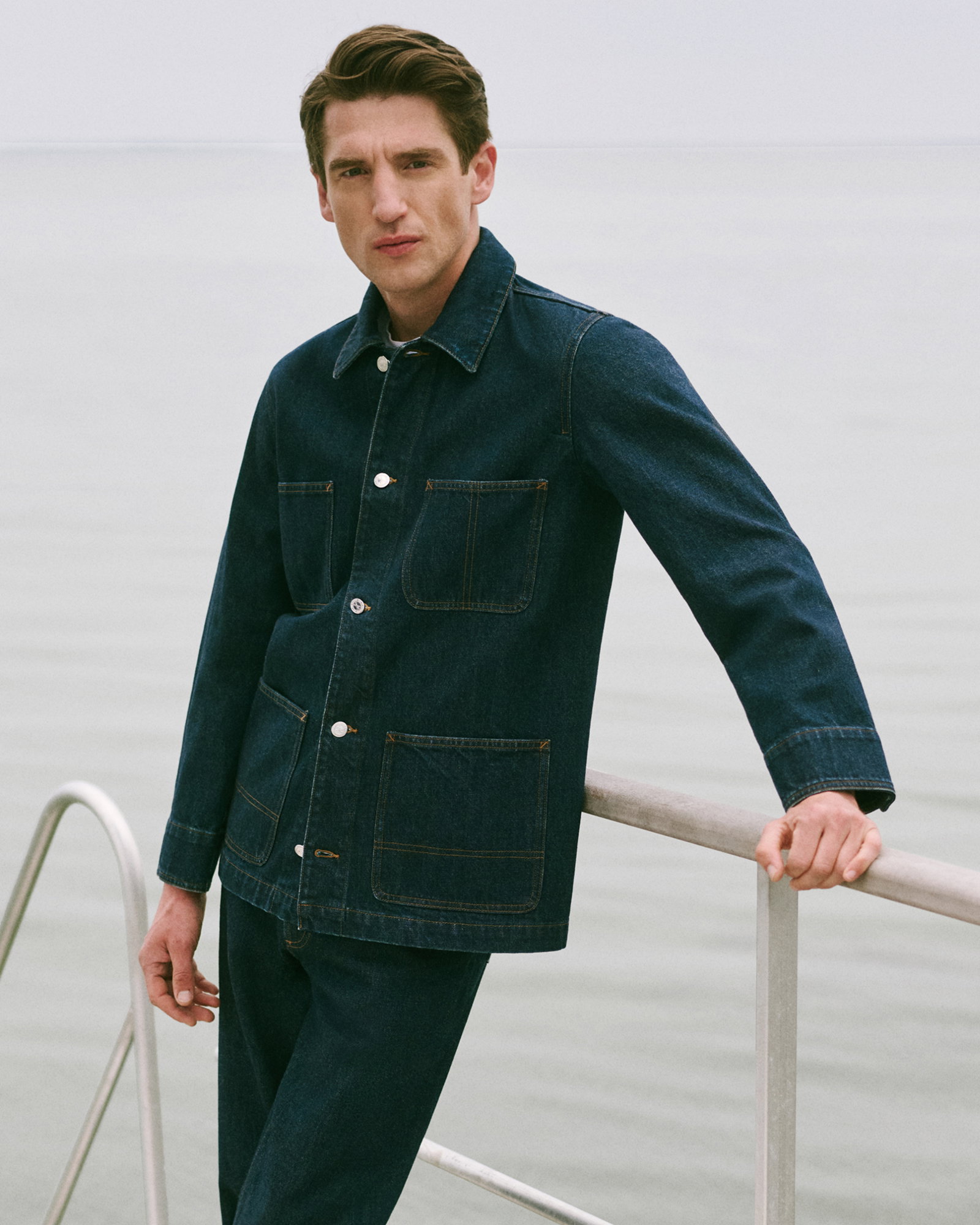 Mann lehnt an Brüstung vor See, trägt dunkelblaue Jeansjacke und Jeanshose, hat braune Haare und ernsten Gesichtsausdruck.
