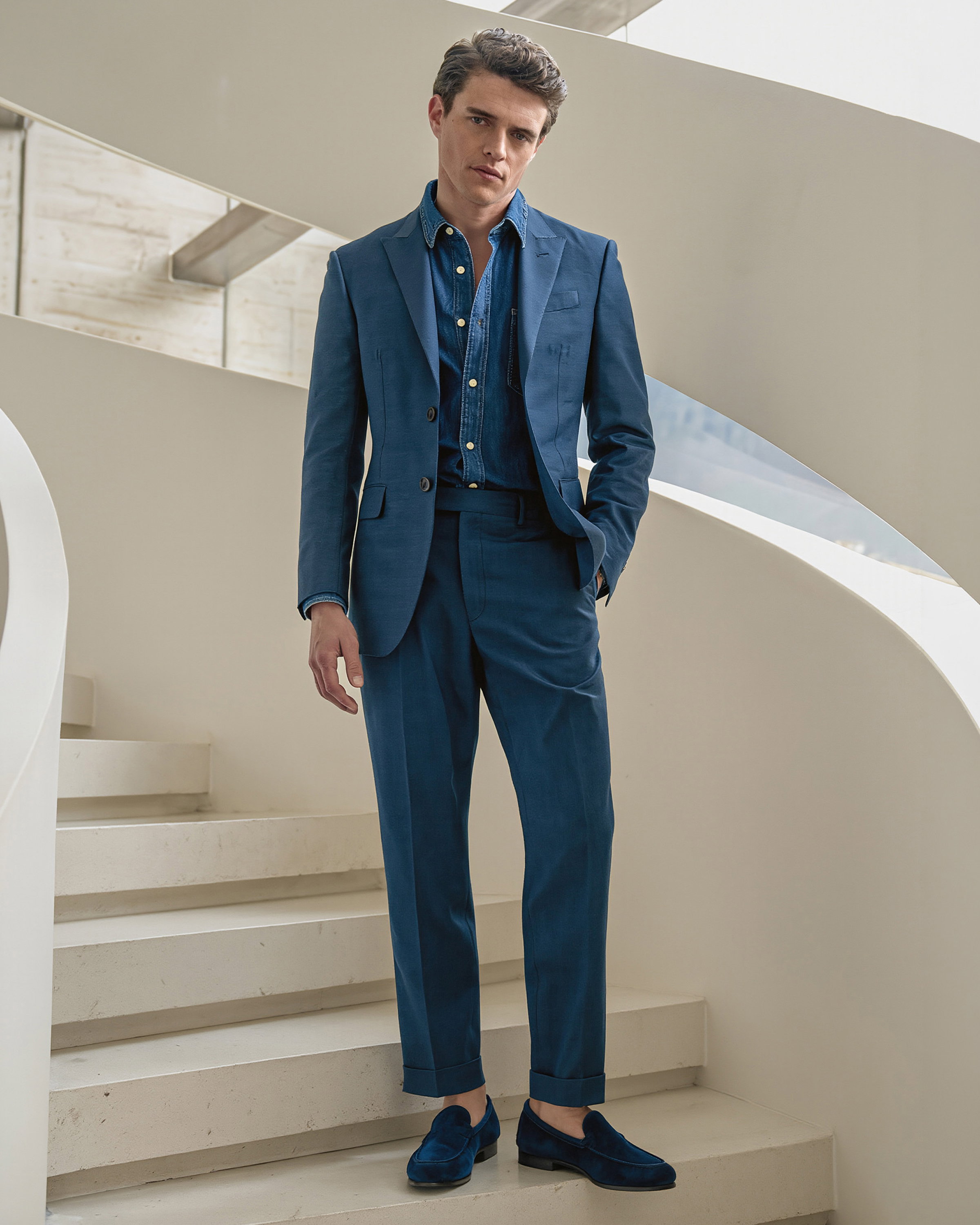 Mann steht auf weißer Treppe, trägt marineblauen Anzug, blaues Jeanshemd und dunkelblaue Loafer, moderner Stil.